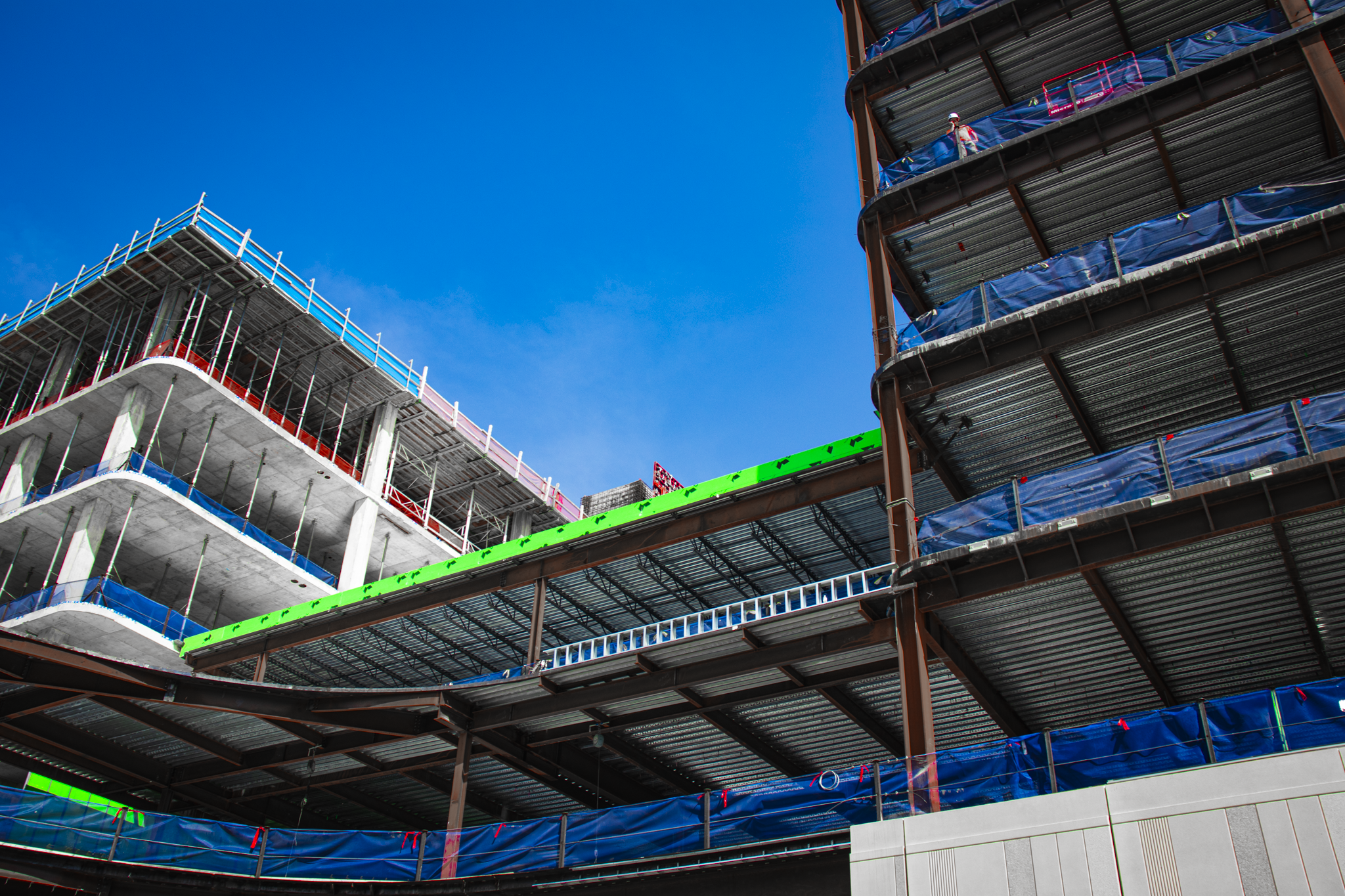 Construction site with multi-story building framework, safety cones, a stop sign, and warning signs, with cars parked nearby under a blue sky with clouds.