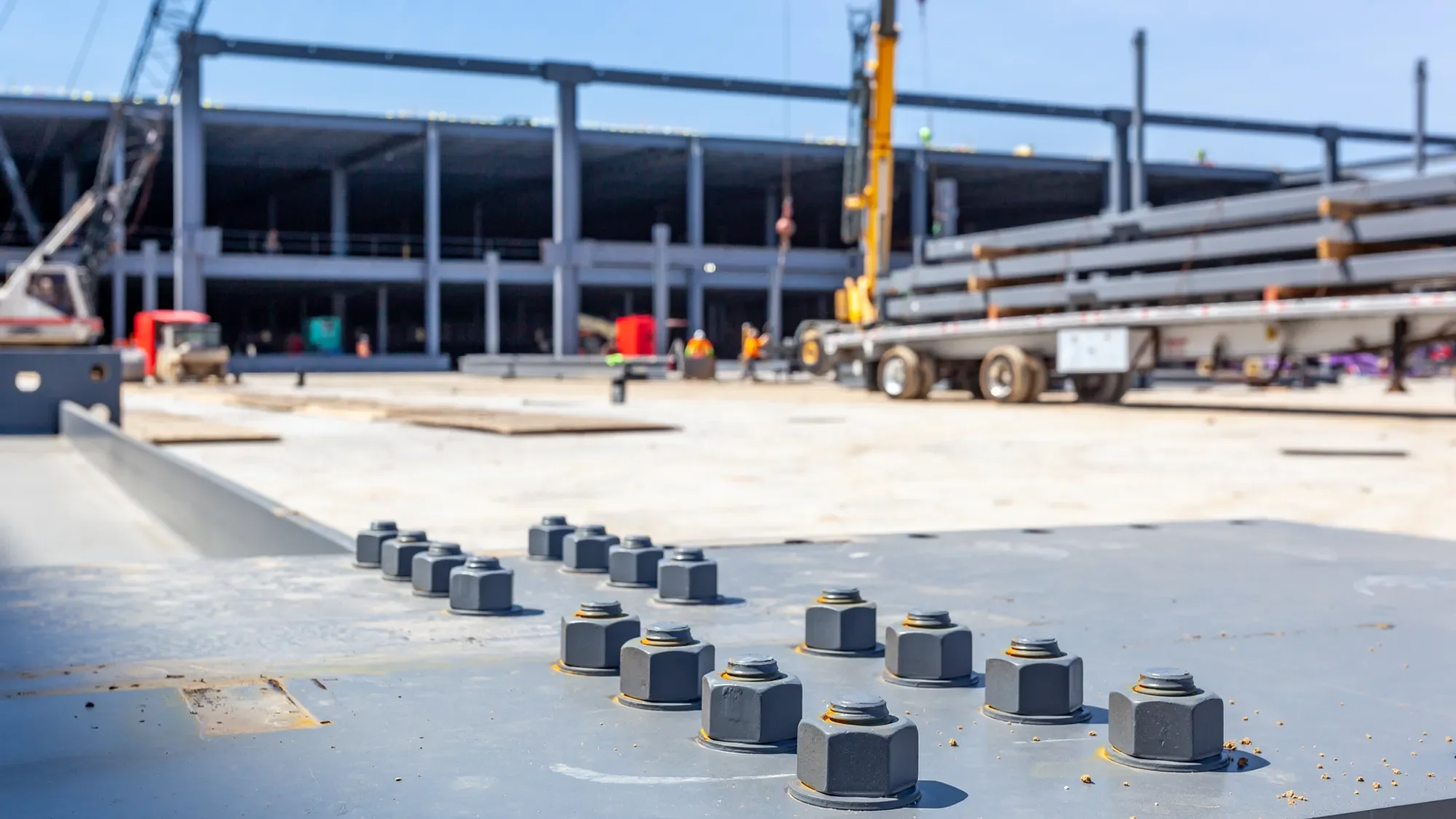 Close-up of a construction site with a metal surface and bolts in the foreground, with a partially constructed building and construction workers in the background.