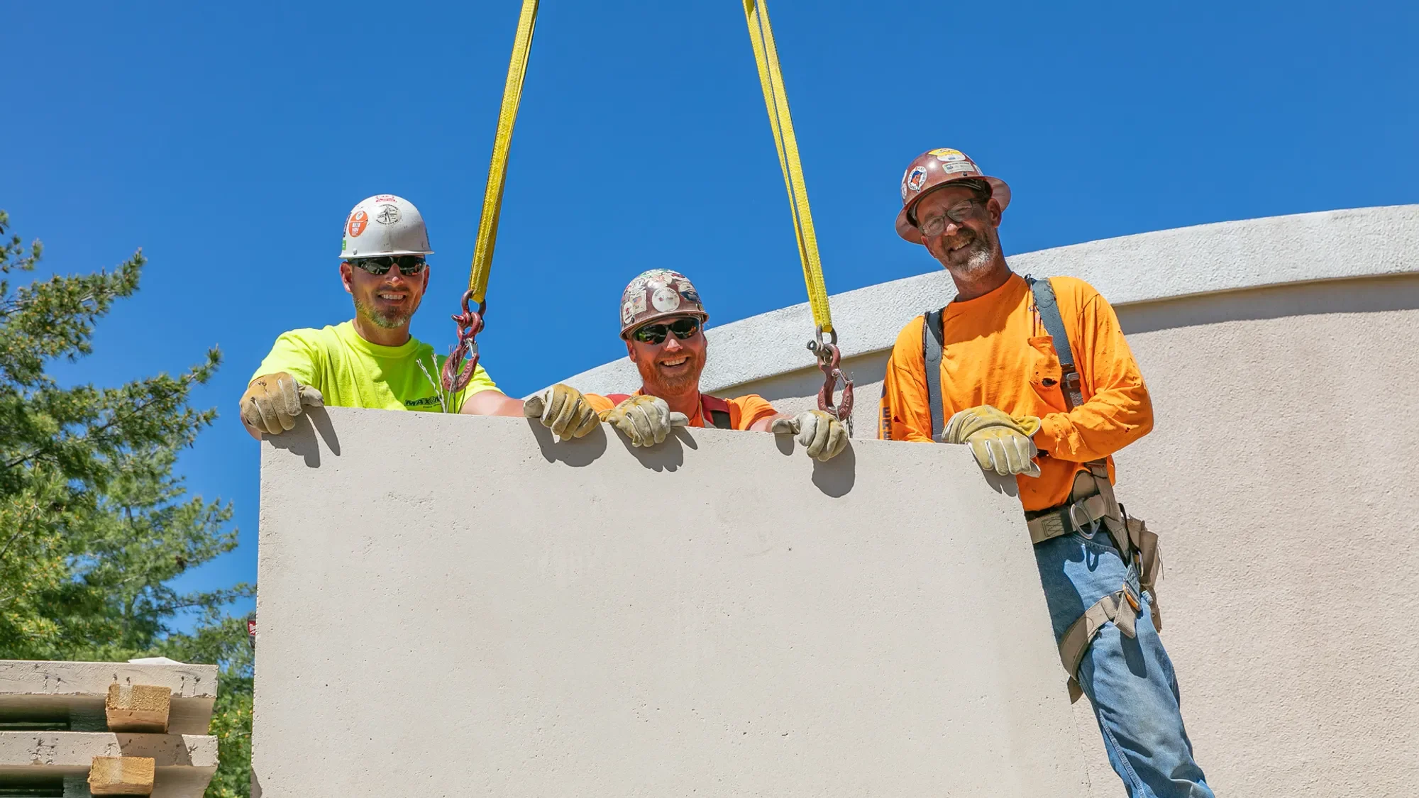 Three construction workers in hard hats and safety gear standing on a building ledge, smiling at the camera with blue sky in background.