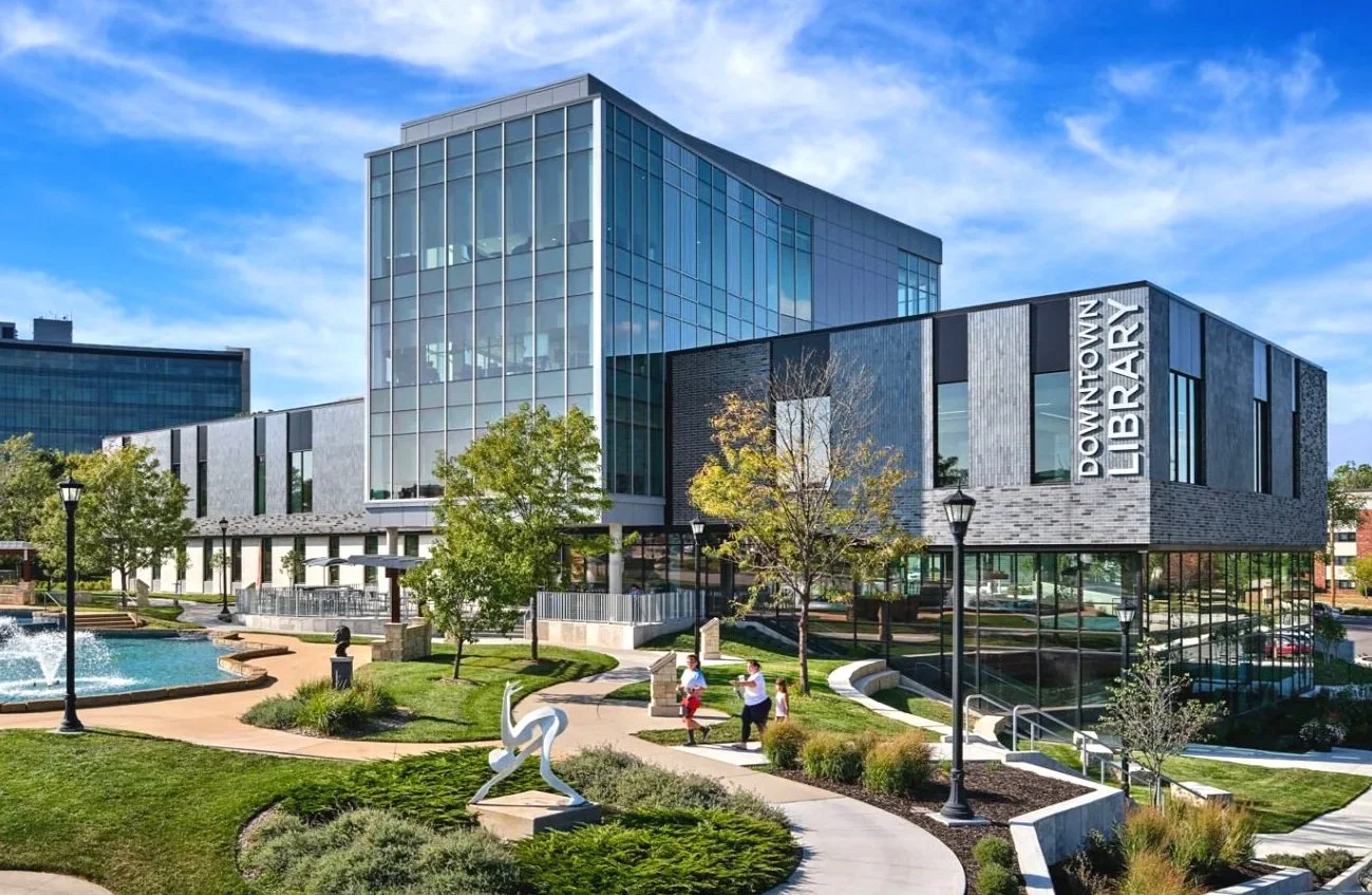 Downtown Library building with glass and modern architecture, trees, and people walking in a park area with grass, pathways, and a fountain.