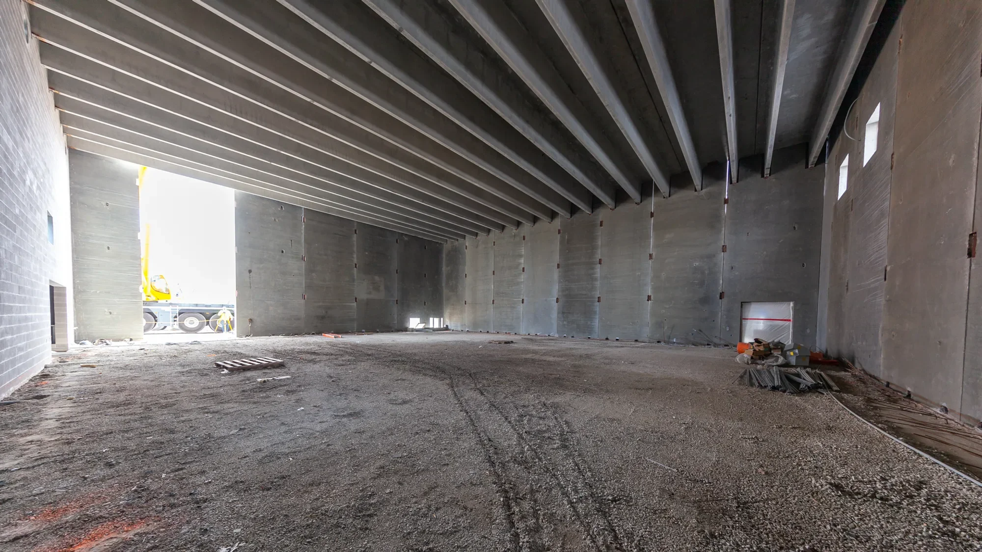 Interior of a building under construction with concrete walls and ceiling, unfinished ground, construction materials, and a view of construction equipment outside.