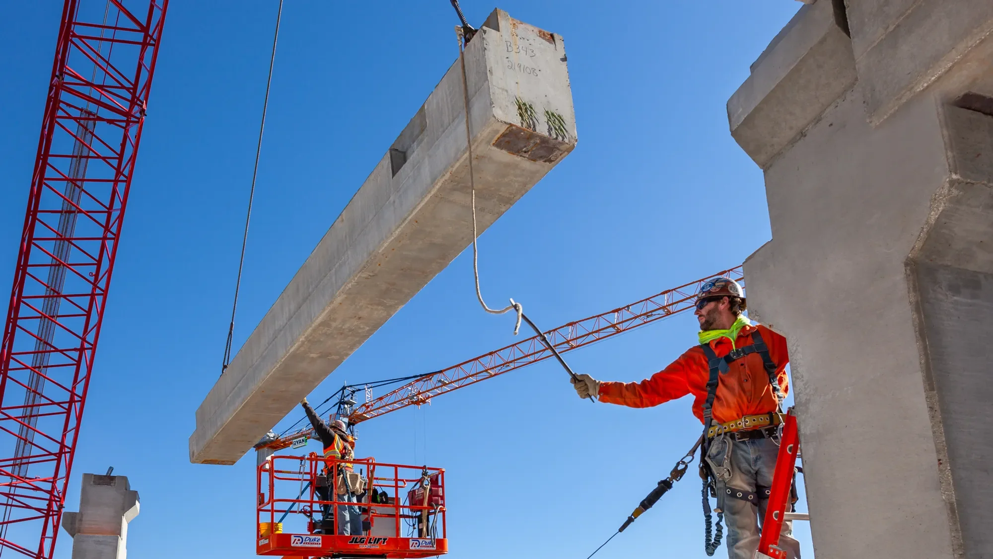 Workers on a construction site guiding a large concrete beam being lifted by a crane into position.
