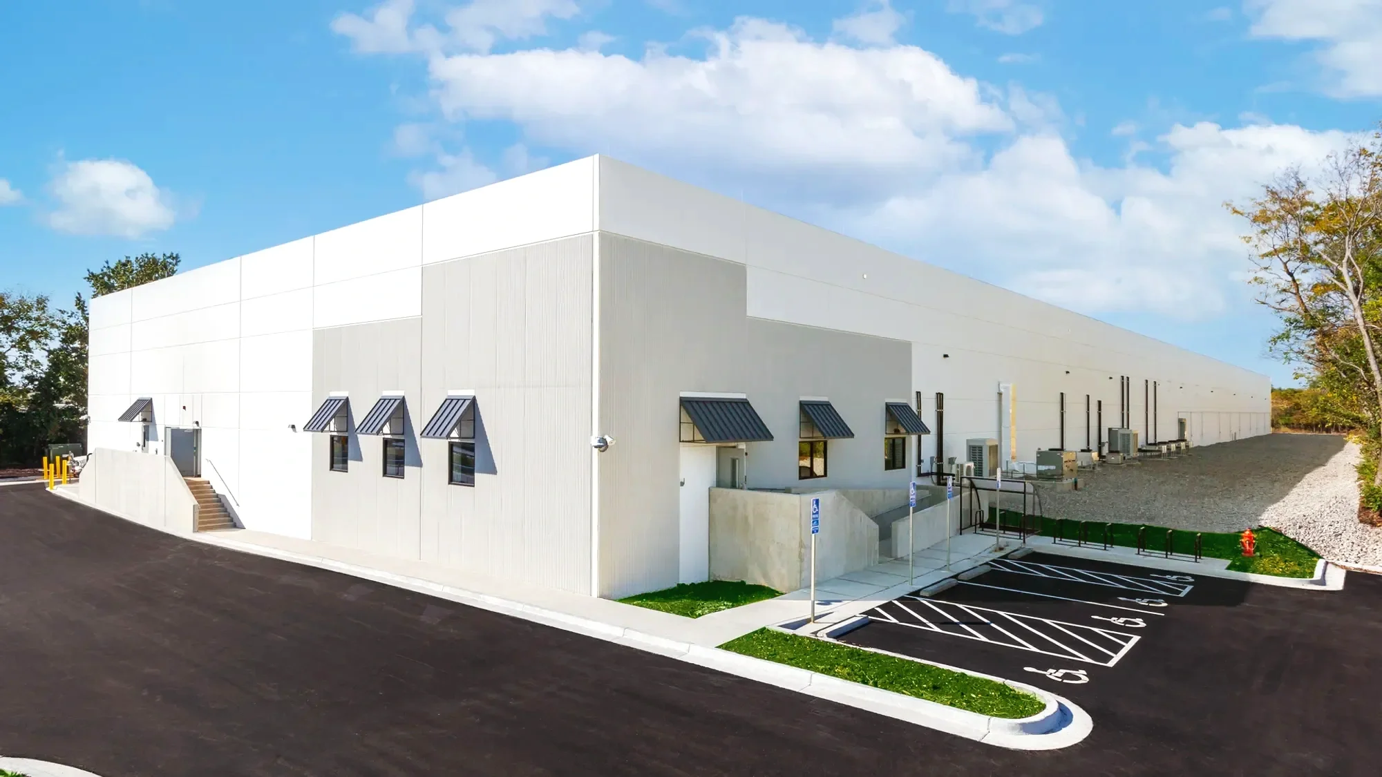 White commercial or industrial building with small awnings over windows, parking spaces with handicap signs, and a driveway, situated in a suburban area with trees and a partly cloudy sky.