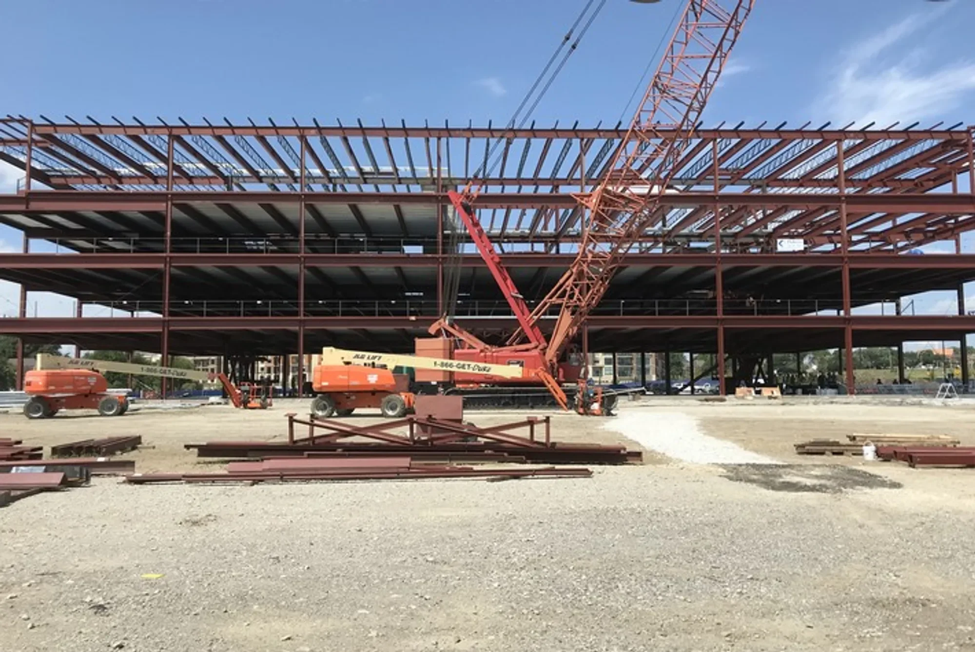 Construction site with a building framework being assembled, orange cranes, construction vehicles, and materials on the ground.
