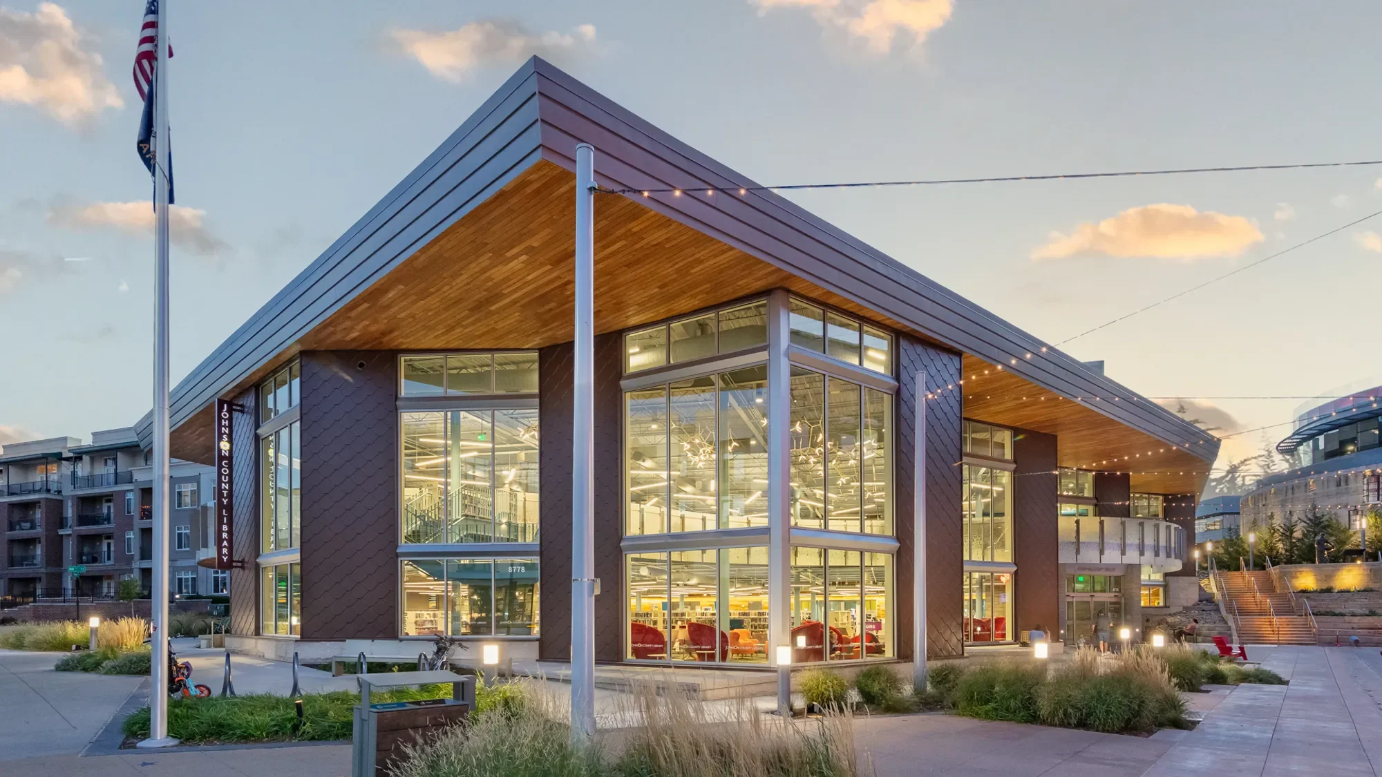 A modern city library building with large glass windows, wooden accents, and cityscape surroundings during dusk, with streetlights and a flagpole in the foreground.