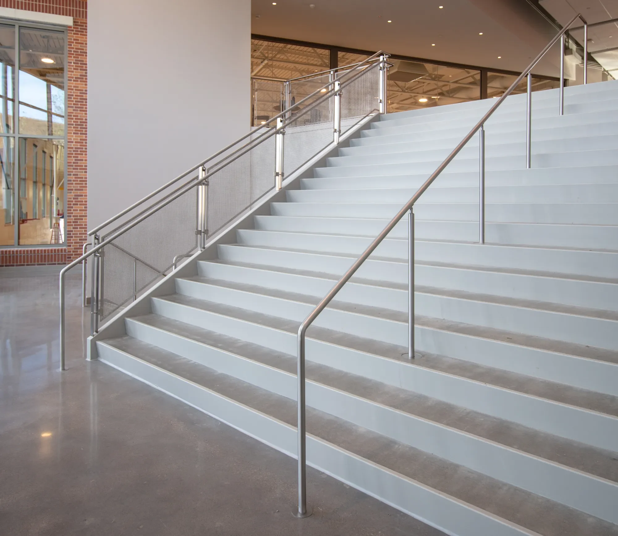 Interior of a building with a wide staircase with white steps and metal handrails, large windows on the left, exposed brick wall, and a modern ceiling with recessed lighting.