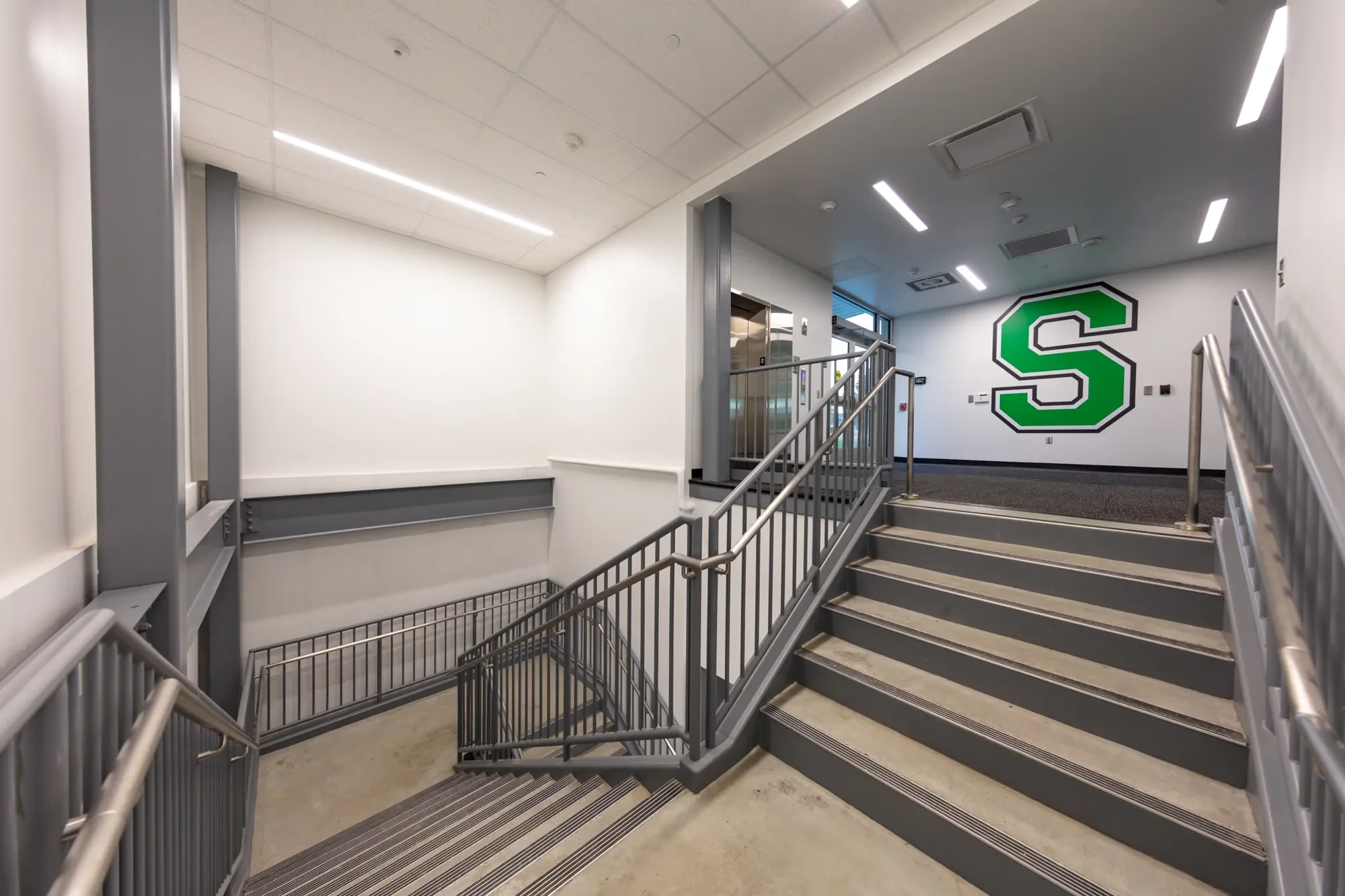 Interior of a modern building featuring a staircase with gray metal railings, an elevator, and a white wall with a large green letter 'S' outlined in black.