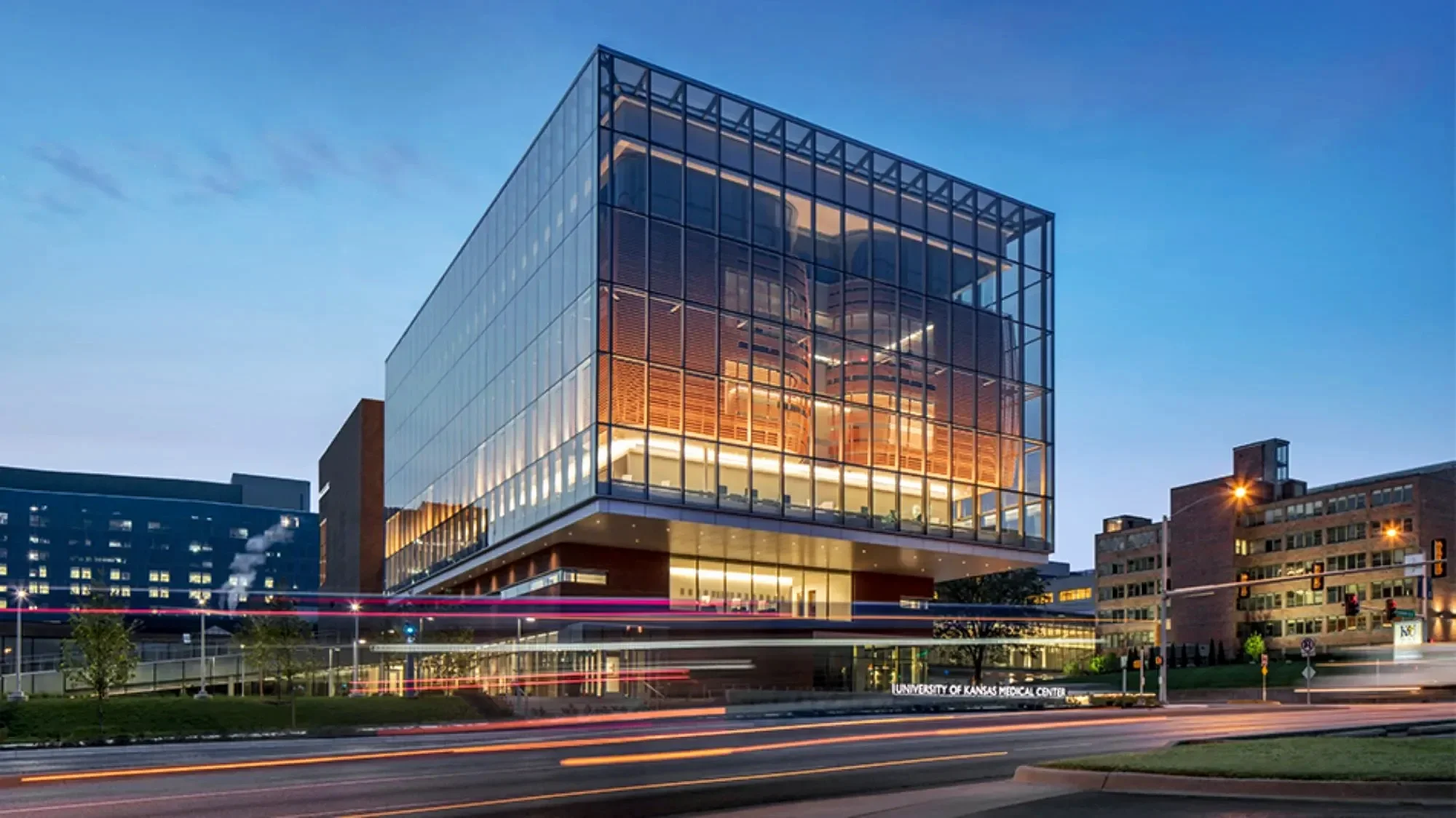 Modern glass building with illuminated interior, street with light trails from passing cars, and a sign indicating University of Kansas Medical Center