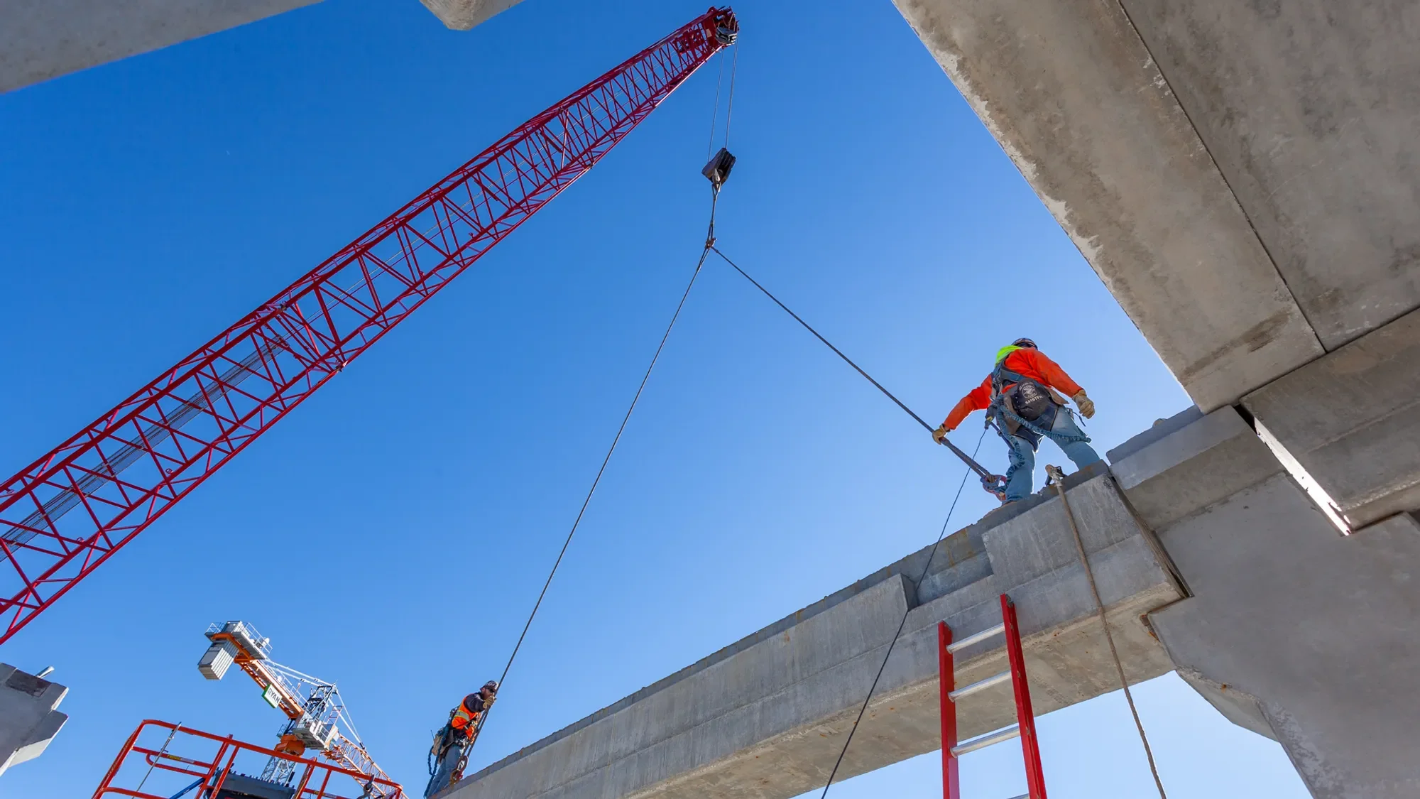 Construction workers on a concrete structure working on a building under a clear blue sky, with a large red crane overhead.