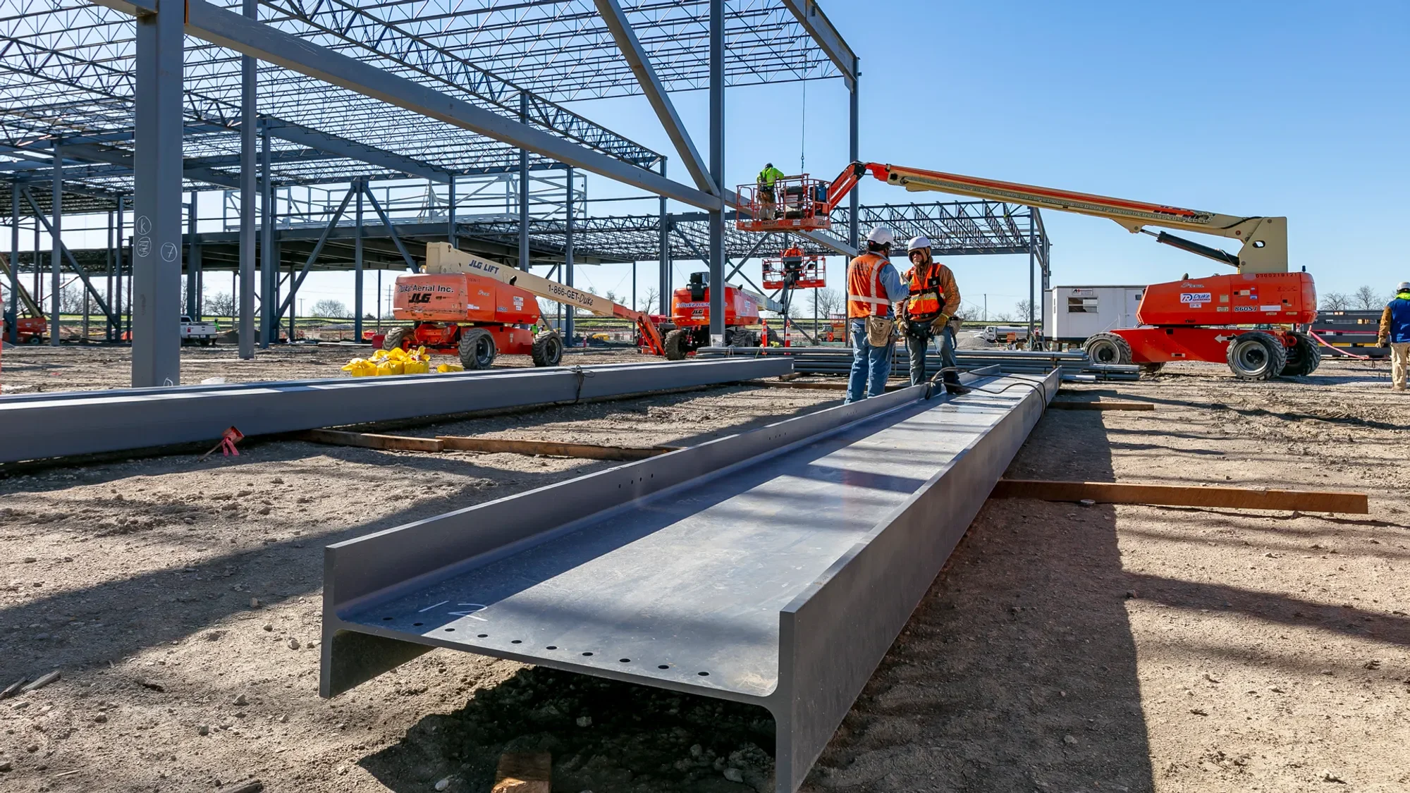 Construction workers are building a large metal structure outdoors, using cranes and bucket lifts. Metal beams are laid out on the ground and some are being assembled into the framework of a building.