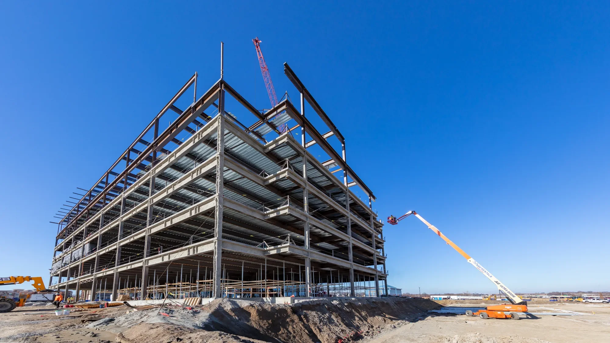 A multi-story building under construction with a crane and construction workers on site beneath a clear blue sky.
