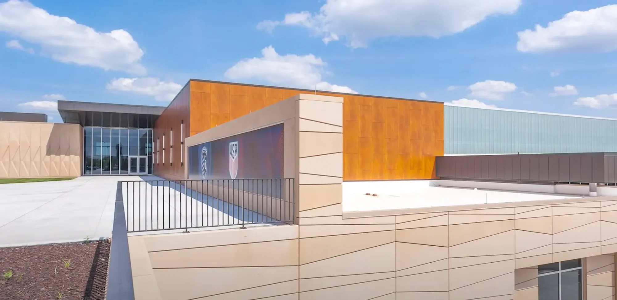 Modern building with orange, beige, and light blue exterior walls, large glass entrance, and an MLS logo sign, under a partly cloudy sky.
