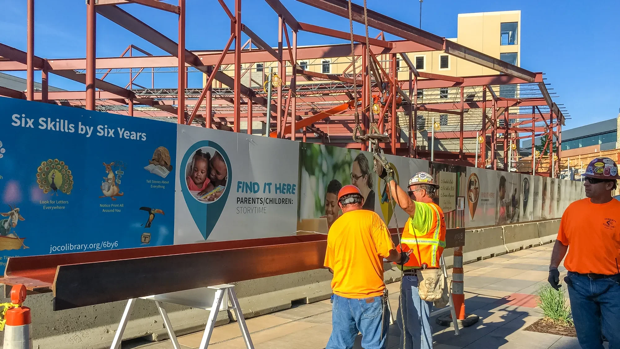 Construction workers in orange and yellow safety vests and hard hats working at a construction site with a steel framework. A banner on the fence features colorful illustrations and text about childhood skills and parent-child storytime activities.