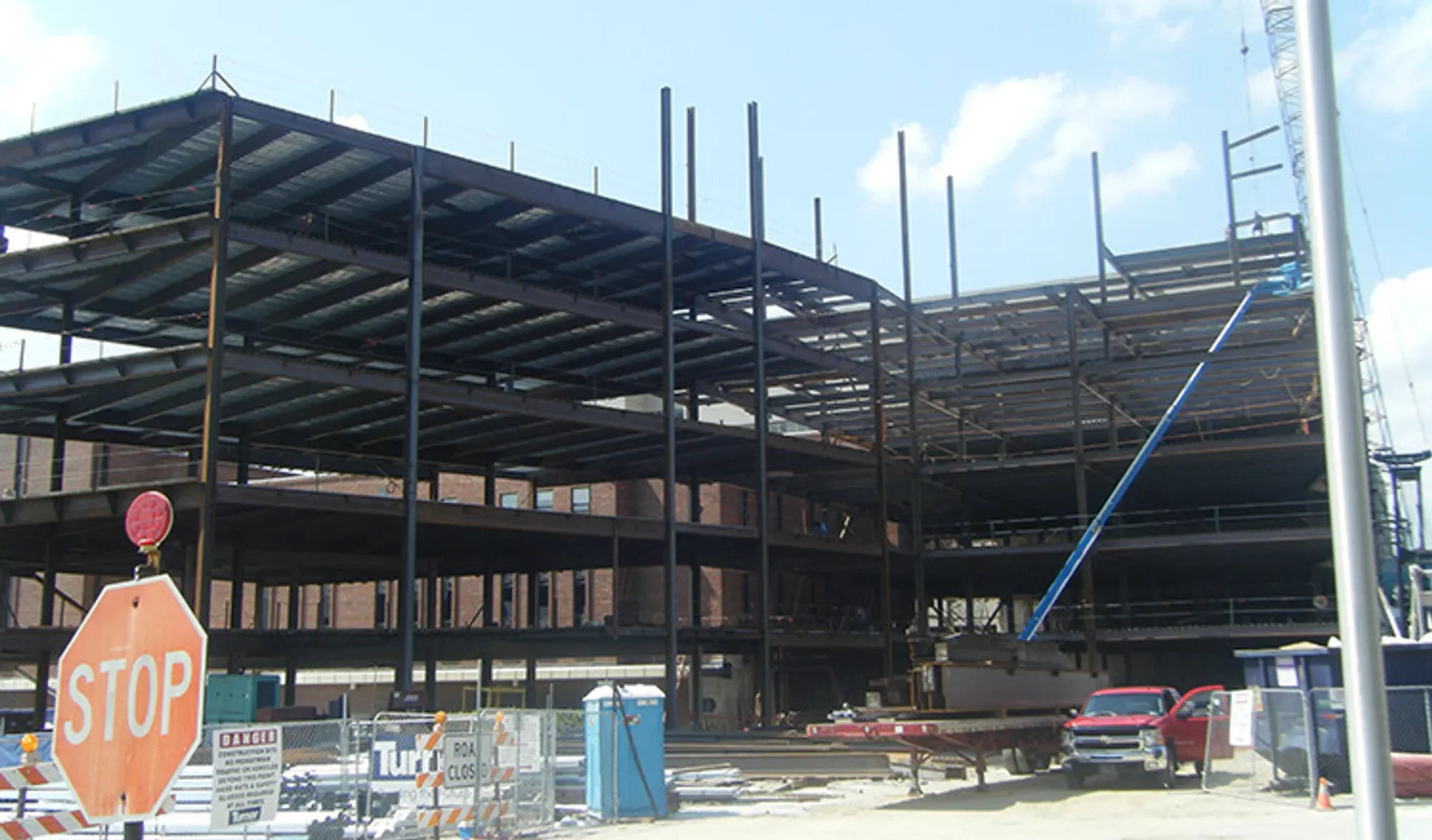 Construction site with multi-story building framework, safety cones, a stop sign, and warning signs, with cars parked nearby under a blue sky with clouds.