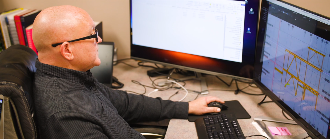 A man with glasses working at a desk with multiple monitors displaying technical drawings and spreadsheets.