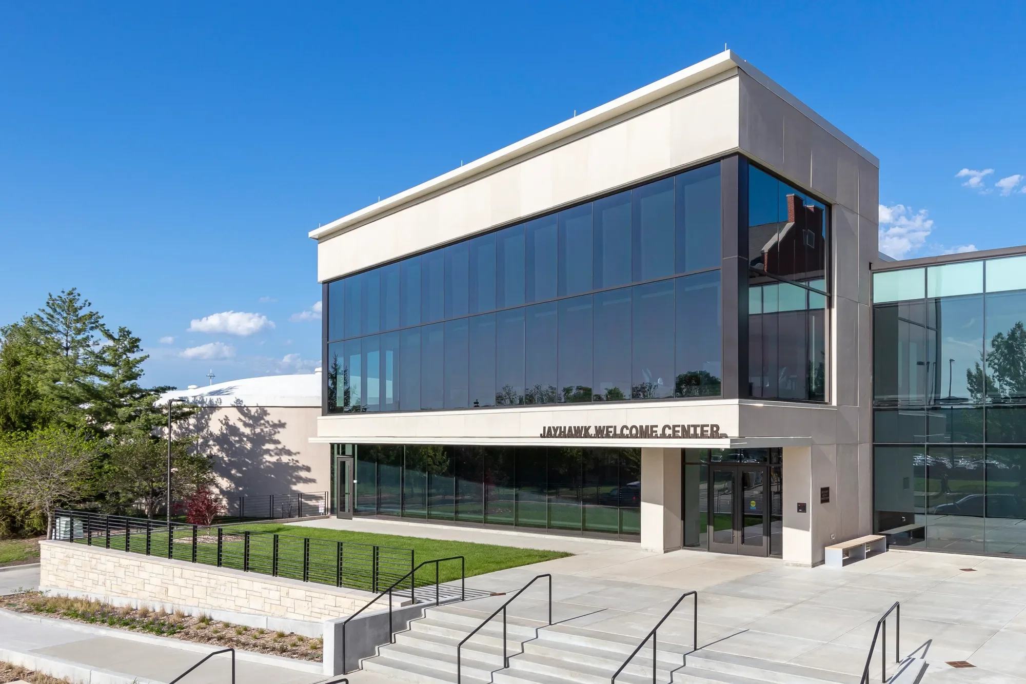 Front view of a modern building with glass windows and a white exterior, labeled 'JAYHAWK WELCOME CENTER', with steps leading up to the entrance, greenery on the side, and a clear blue sky overhead.