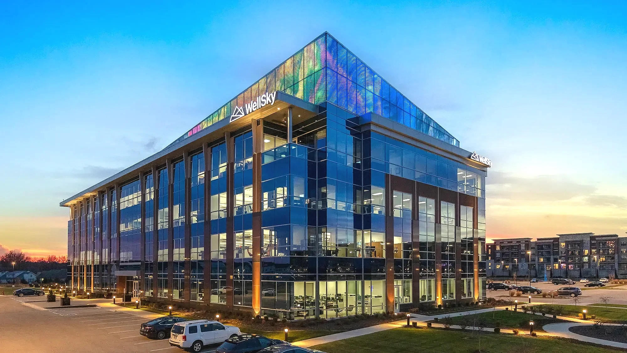 A modern glass office building with colorful illuminated panels at the top, during sunset.