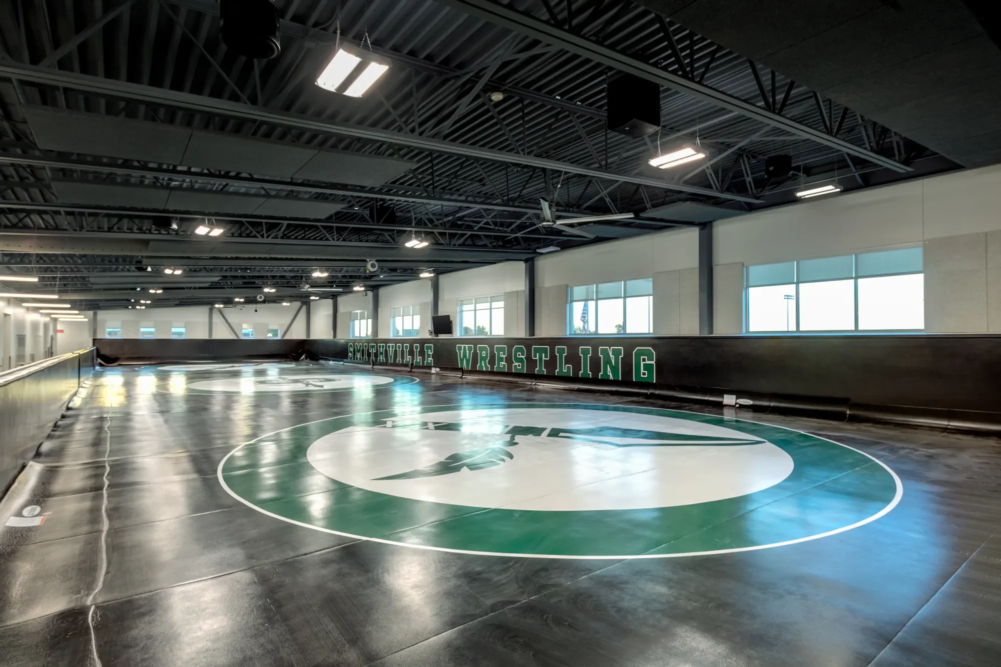 Indoor wrestling mat area with a green and white circle, in a sports facility with large windows and a sign that reads 'Smithville Wrestling' on the wall.