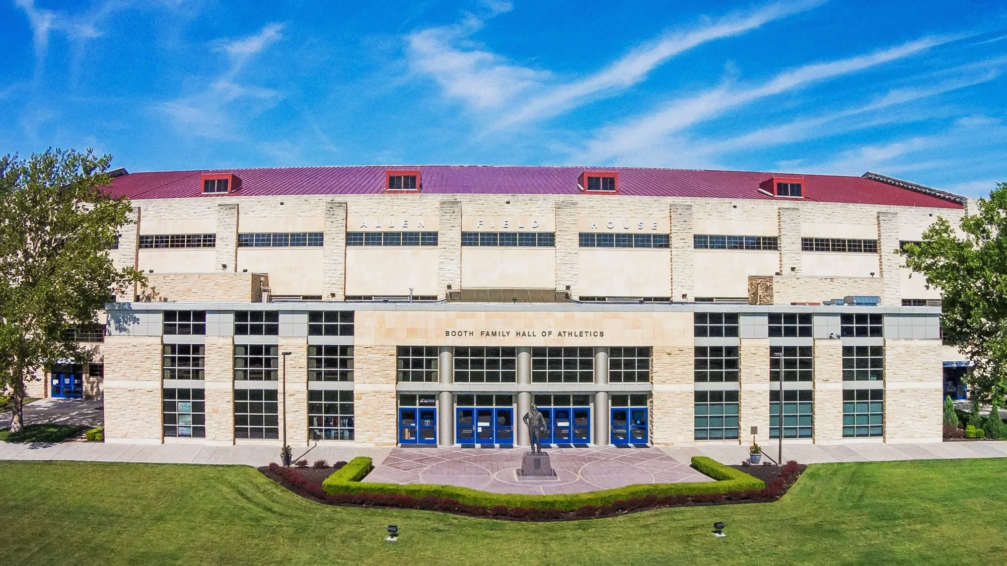 The Allen Field House, a large, modern building with a stone facade, glass windows, and blue doors, is situated under a bright blue sky with some clouds. There is a well-maintained front lawn with grass, small bushes, and two trees. A statue of a person stands in front of the building, and a sign reads 'Booth Family Hall of Athletics' above the entrance.