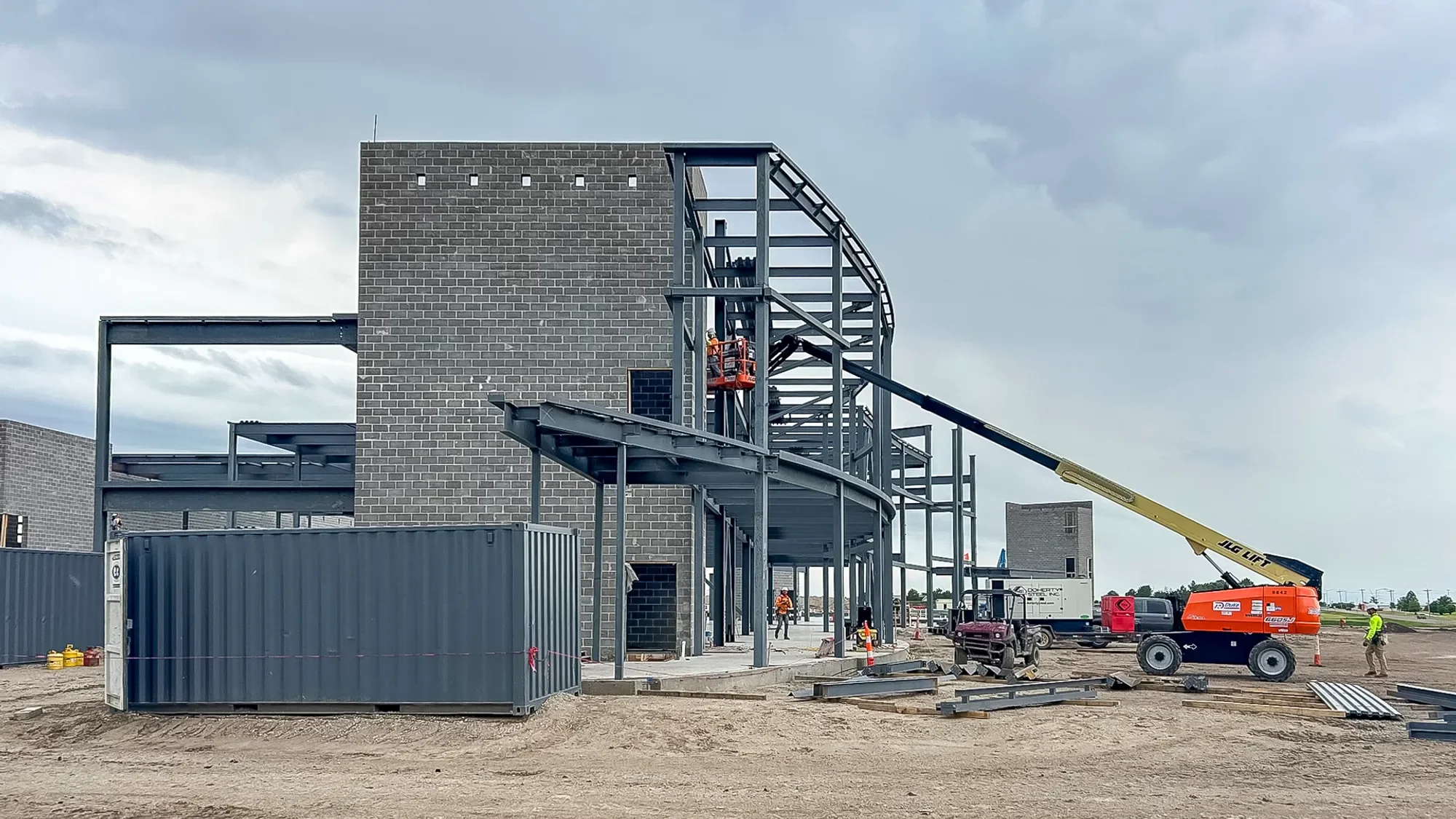 Construction site with steel framework and brick walls, workers in safety gear, a crane lifting materials, and overcast sky.