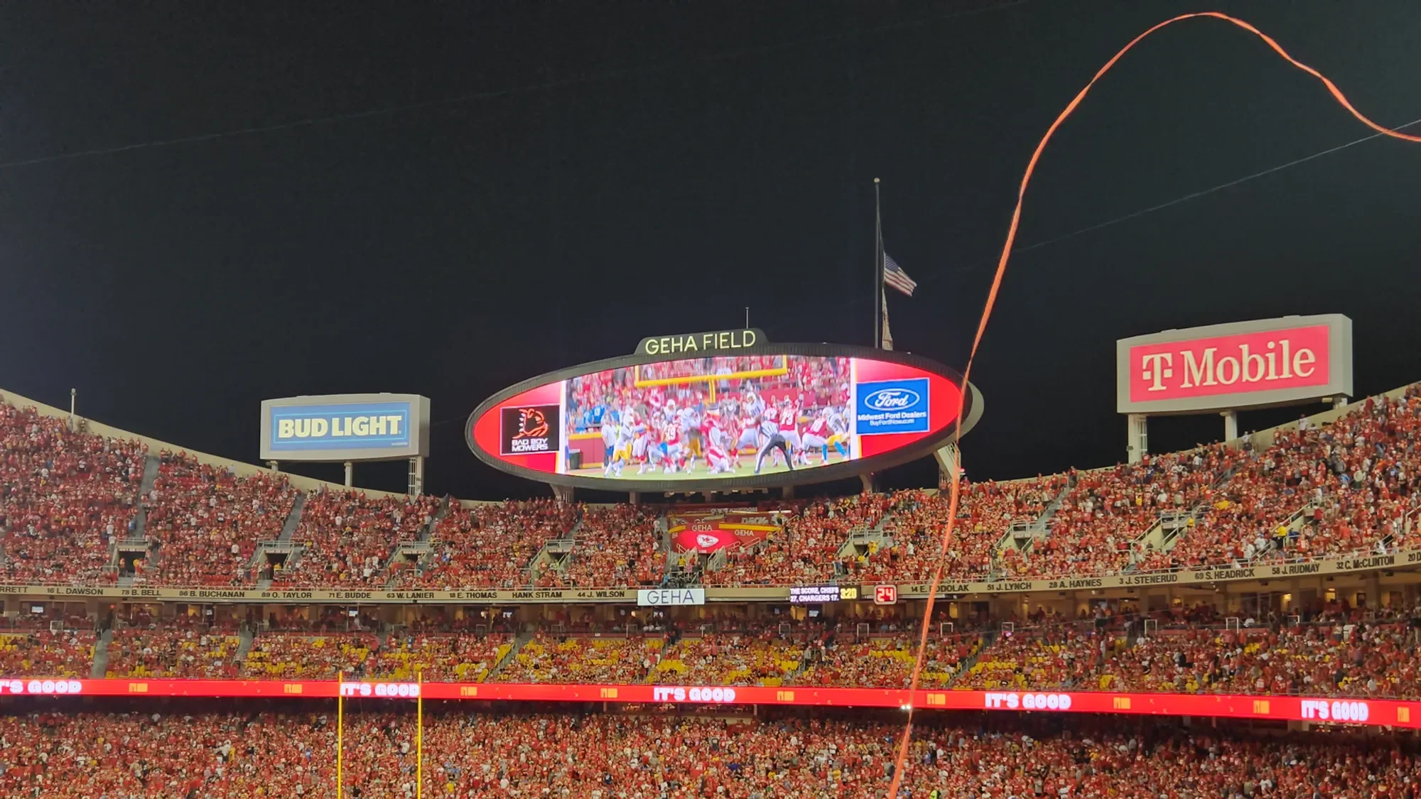 Nighttime view of a crowded football stadium with a large digital scoreboard displaying players on the field and spectators. Flags are visible, including an American flag, and advertisements around the stadium include T-Mobile, Bud Light, and Ford.