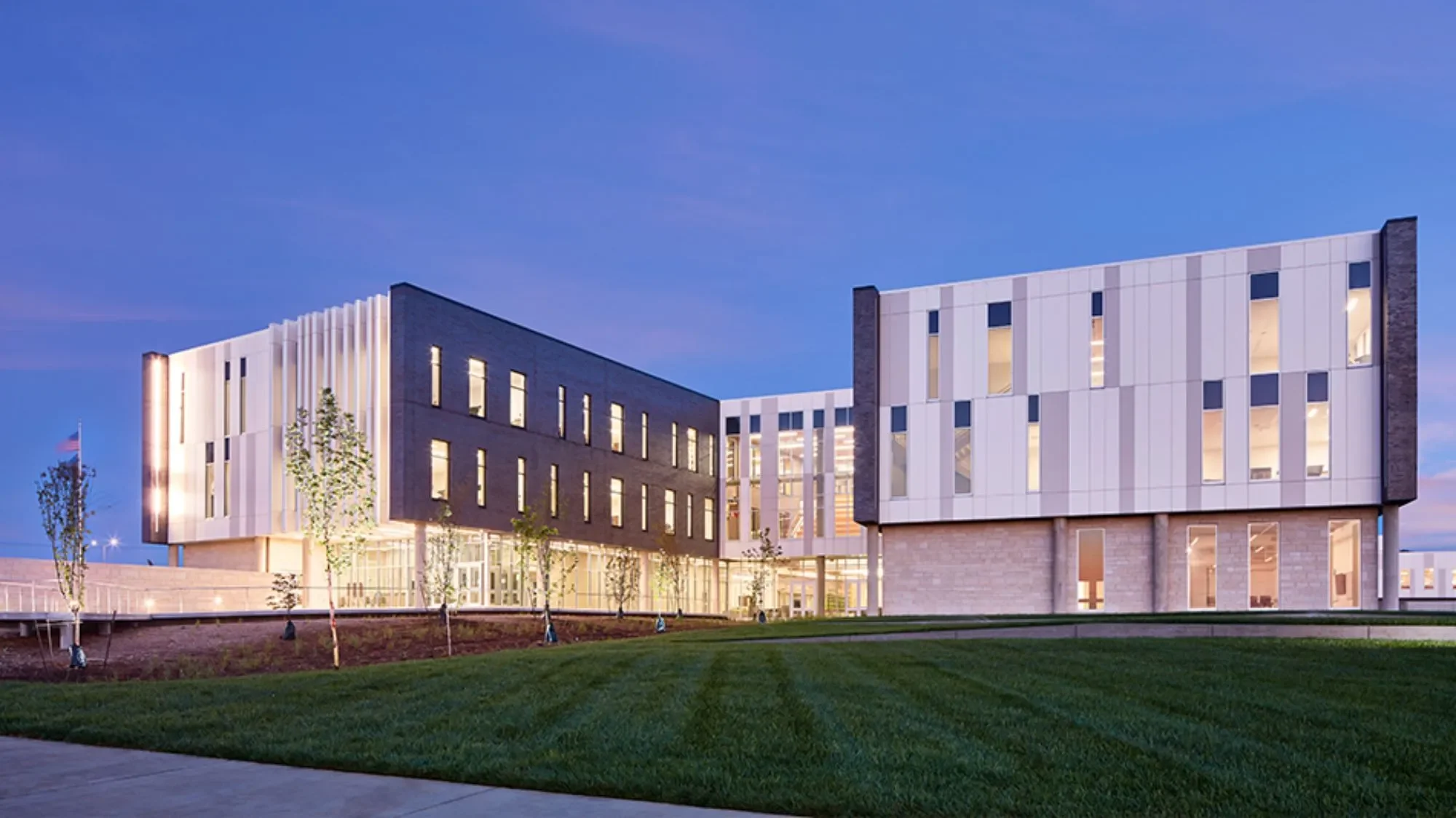 Modern multi-story building with large windows, illuminated from within, set against dusk sky, surrounded by landscaped grass and young trees.