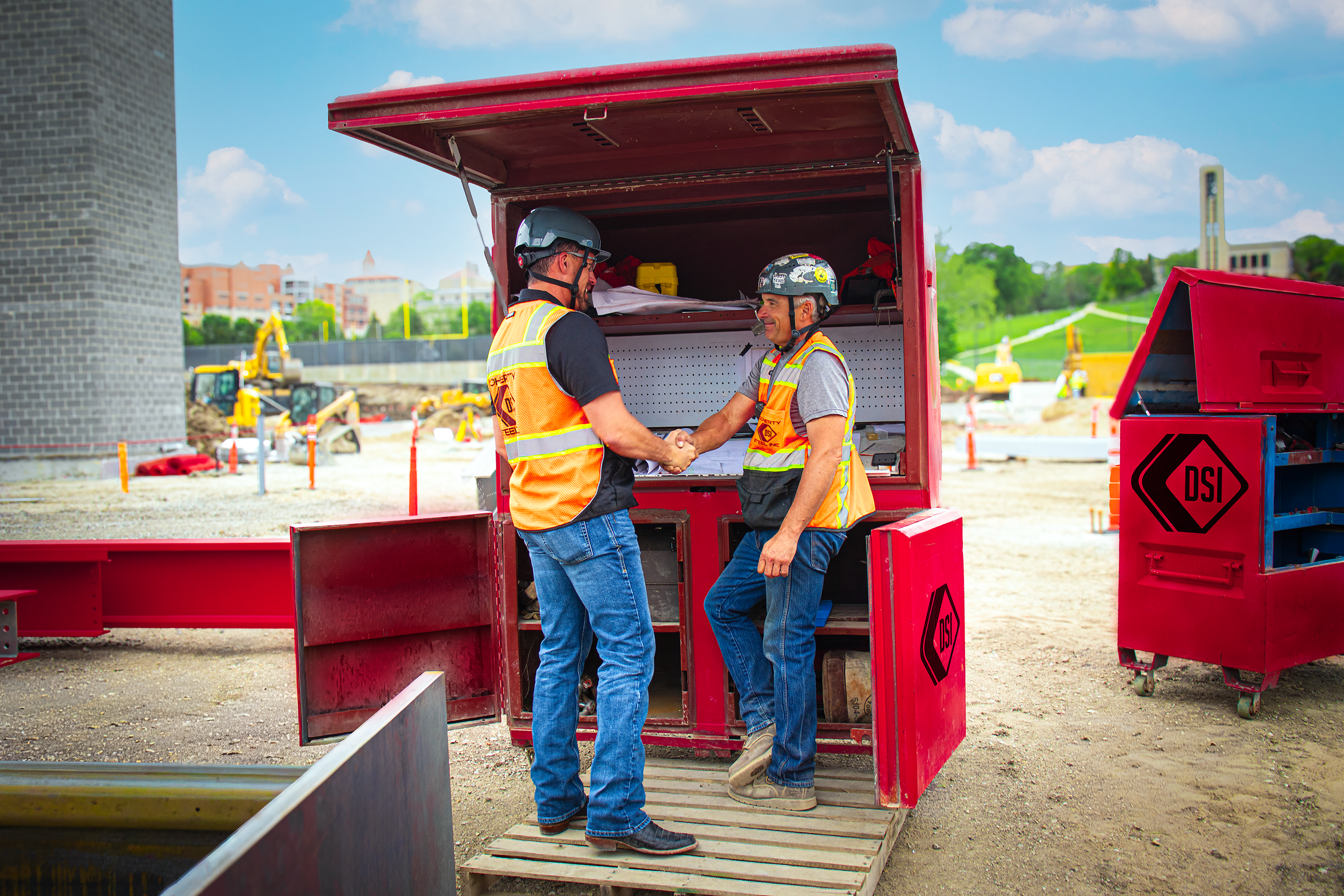Two construction workers shake hands inside a red mobile toolbox on a construction site with machinery and buildings in the background.