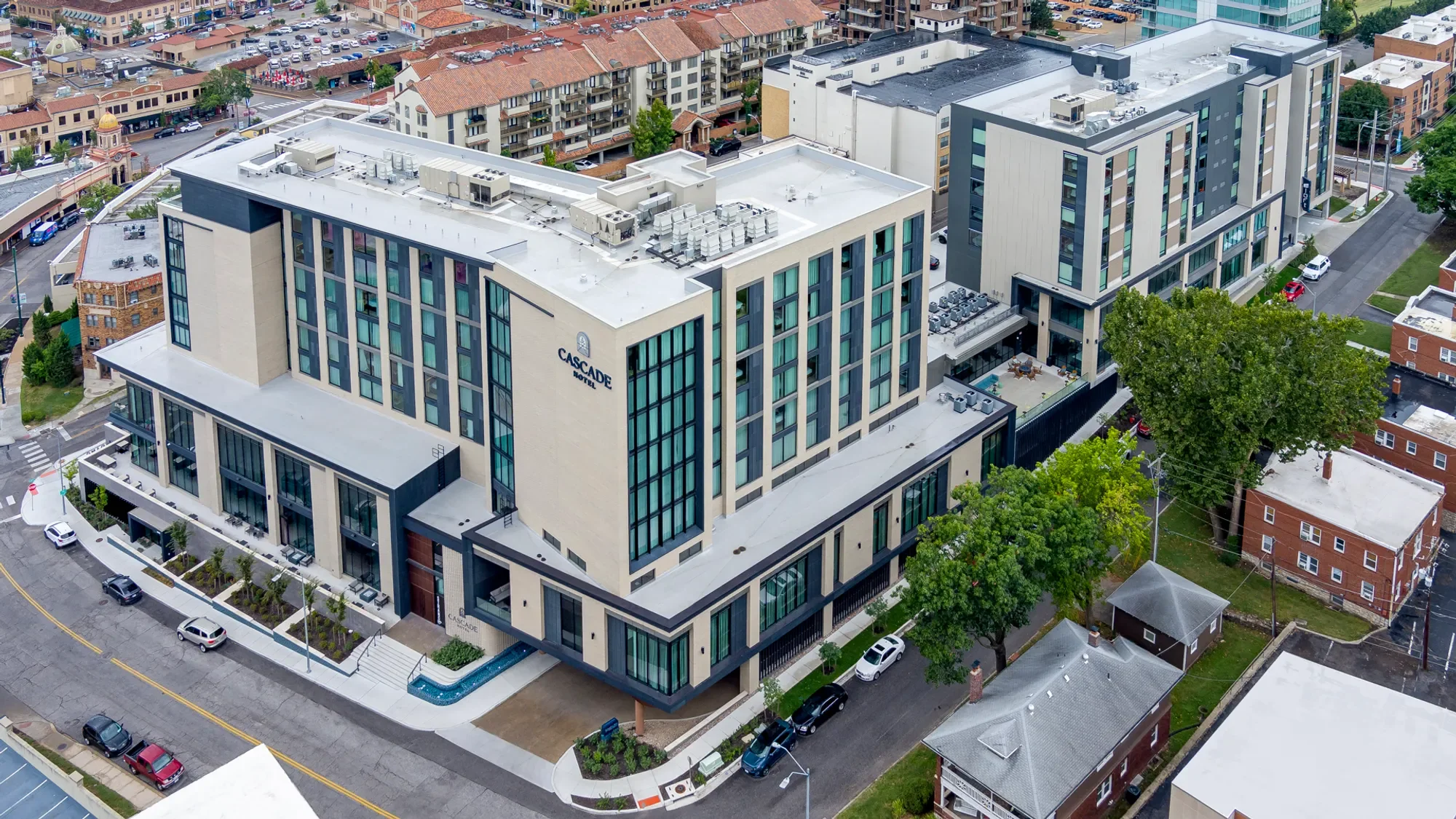Aerial view of a modern hotel with a sign reading 'Cascade Hotel' on the side, surrounded by residential buildings, trees, parked cars, and streets in an urban area.