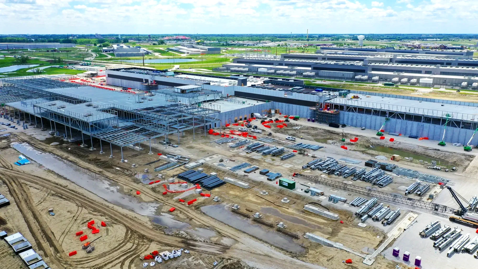 Under construction building with steel framework, construction equipment, and materials, situated in an industrial area with water and green fields in the background.