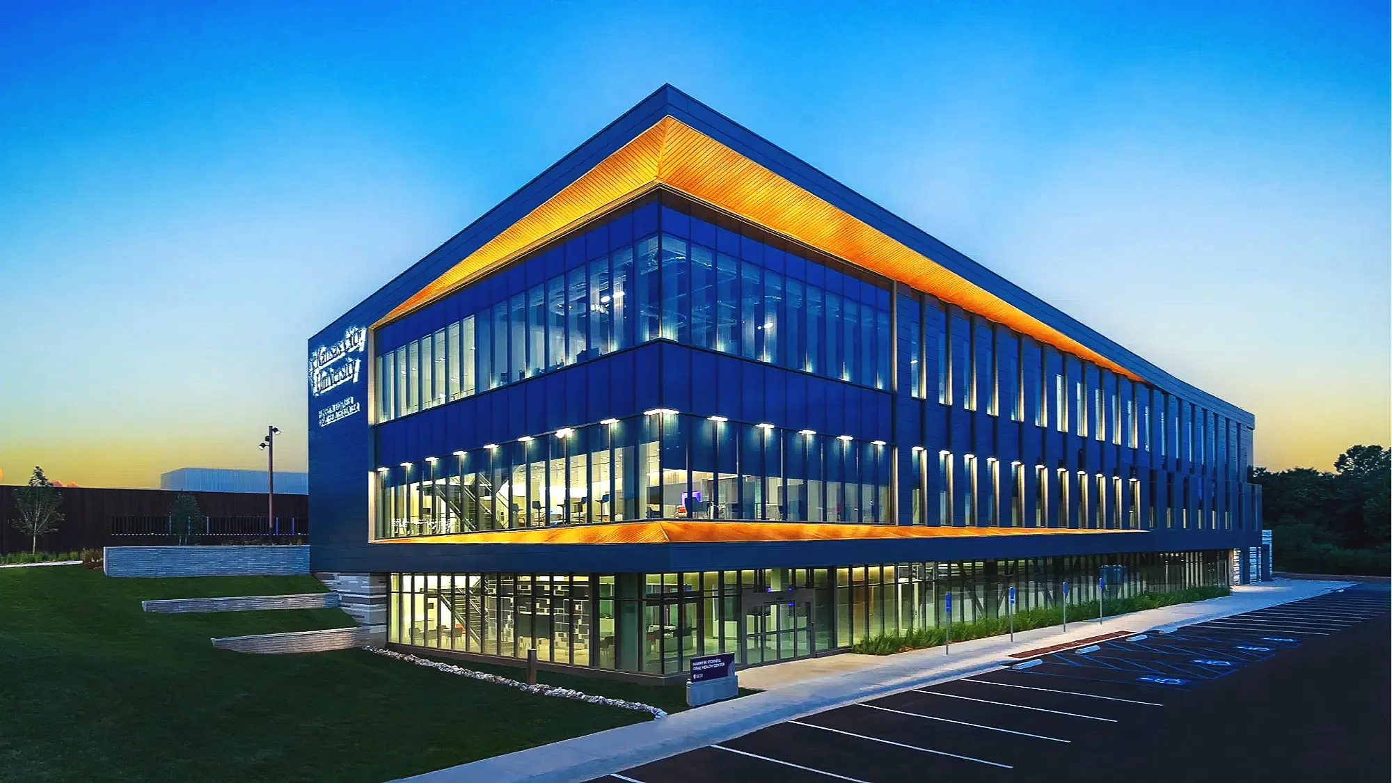 Modern office building with glass facade and yellow accent lighting on the roof edge, taken during dusk with a parking lot in front.