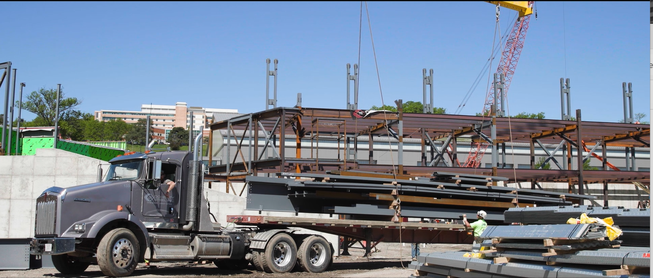 Construction site with a large truck delivering steel beams, workers wearing helmets, and a crane lifting materials against a clear blue sky.