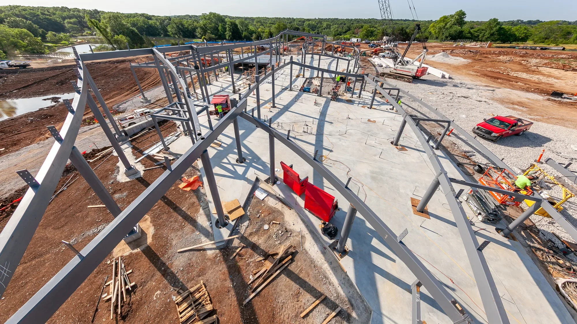Construction workers building a steel framework for a structure on a concrete foundation at a construction site with heavy machinery and trucks nearby.