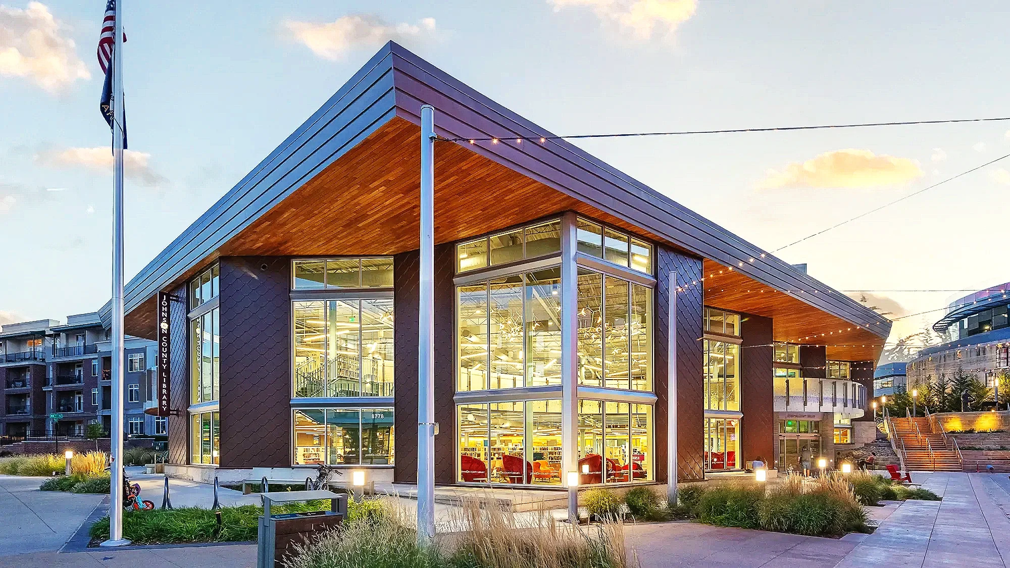Exterior view of a modern library building at dusk with large glass windows, metal and wood accents, flagpole, and surrounding outdoor seating and landscaping.