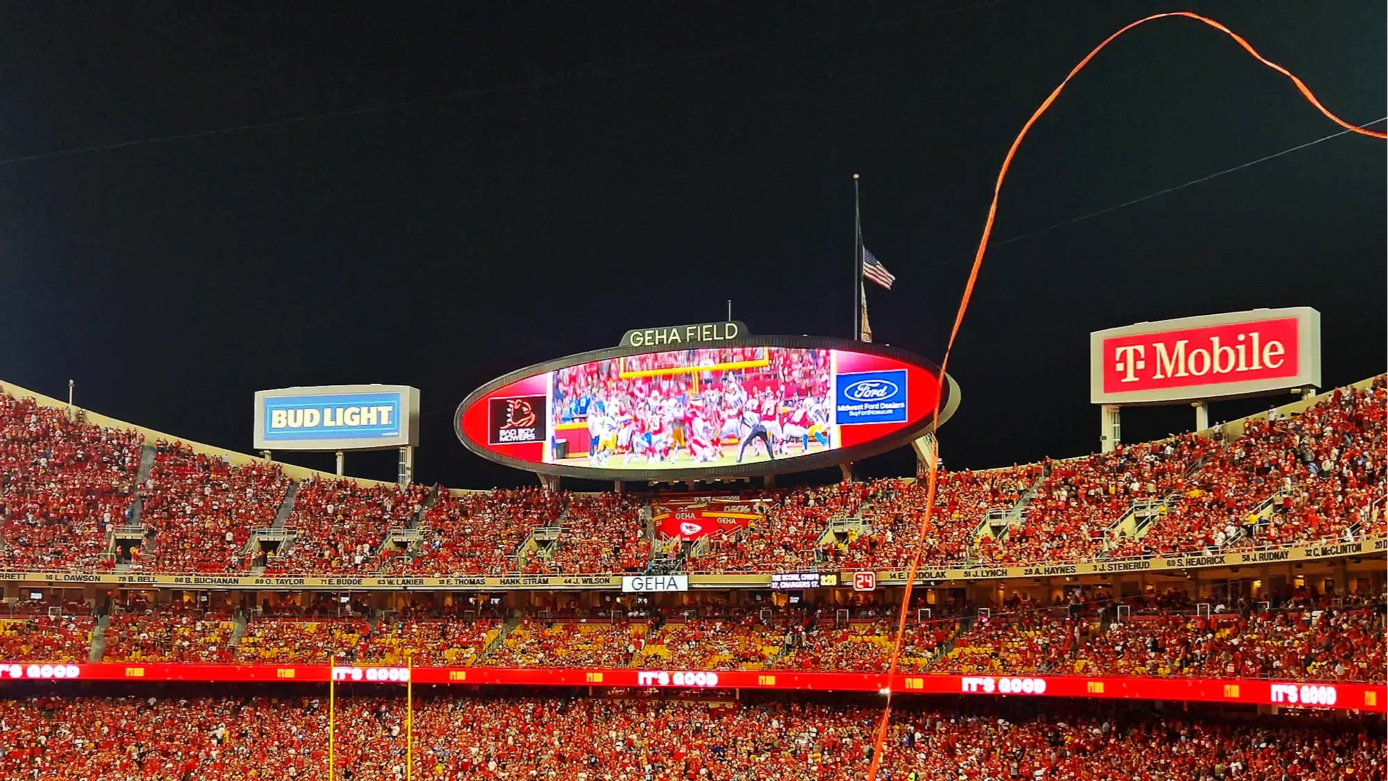 Nighttime scene of the cheifs football stadium filled with spectators, large electronic scoreboard displaying game footage, advertisements for Bud Light, Ford, and T-Mobile, and a visible American flag at the top of the scoreboard.
