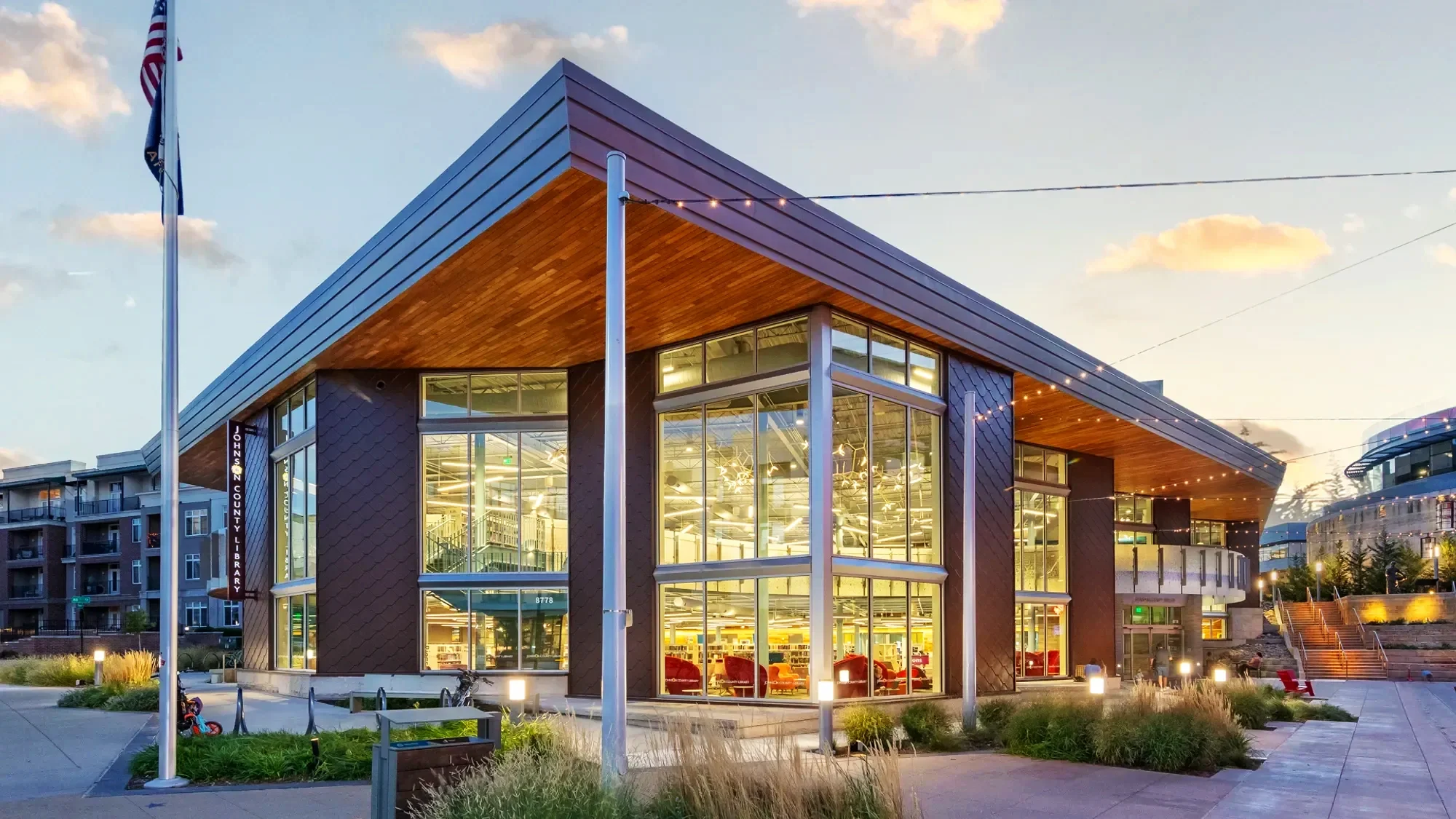 Modern library building with large glass windows and wooden accents, surrounded by outdoor seating and landscaping, during evening with sunset skies.