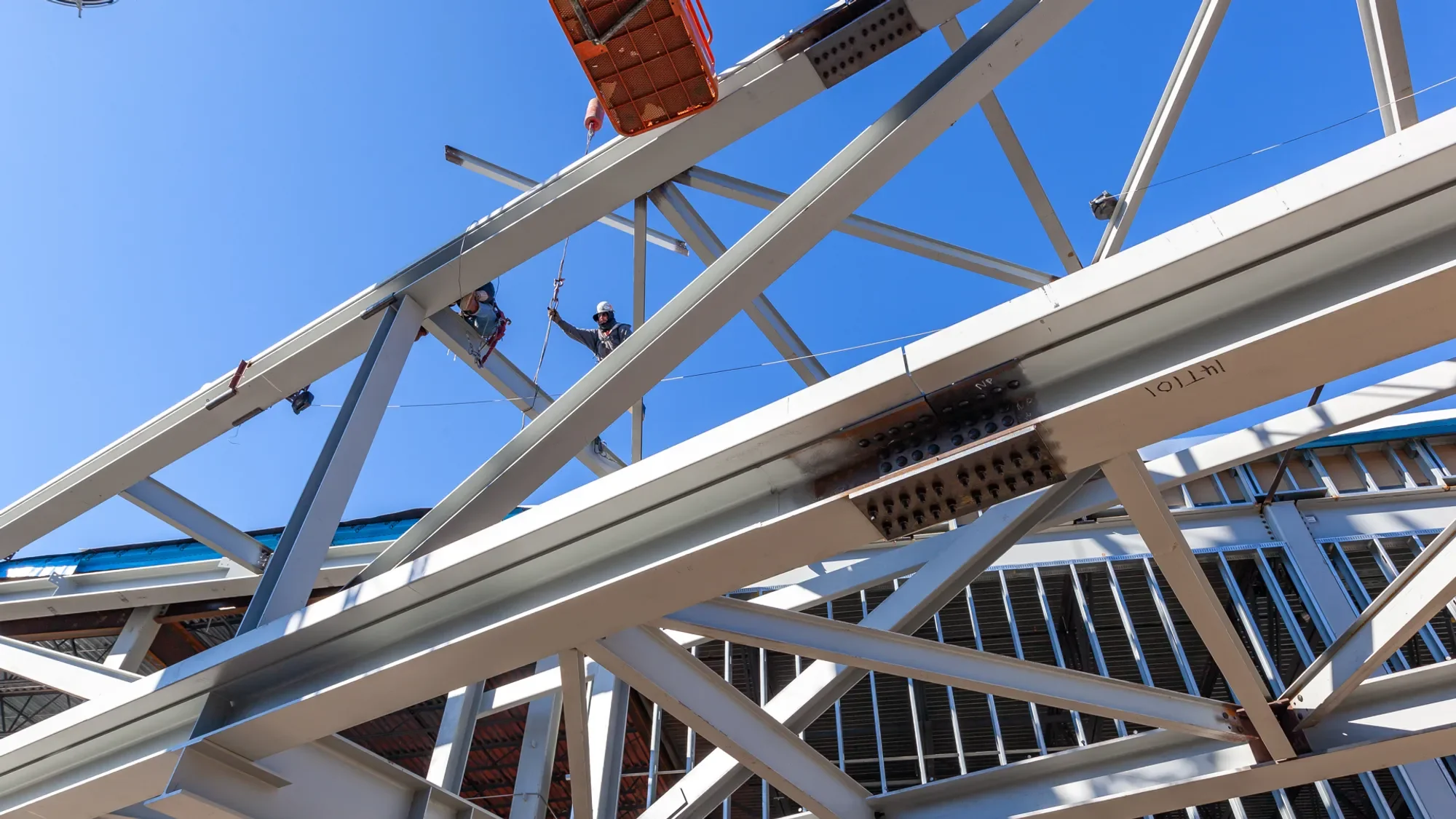 Construction workers wearing safety helmets and harnesses working on the steel framework of a large building under construction, seen against a clear blue sky.