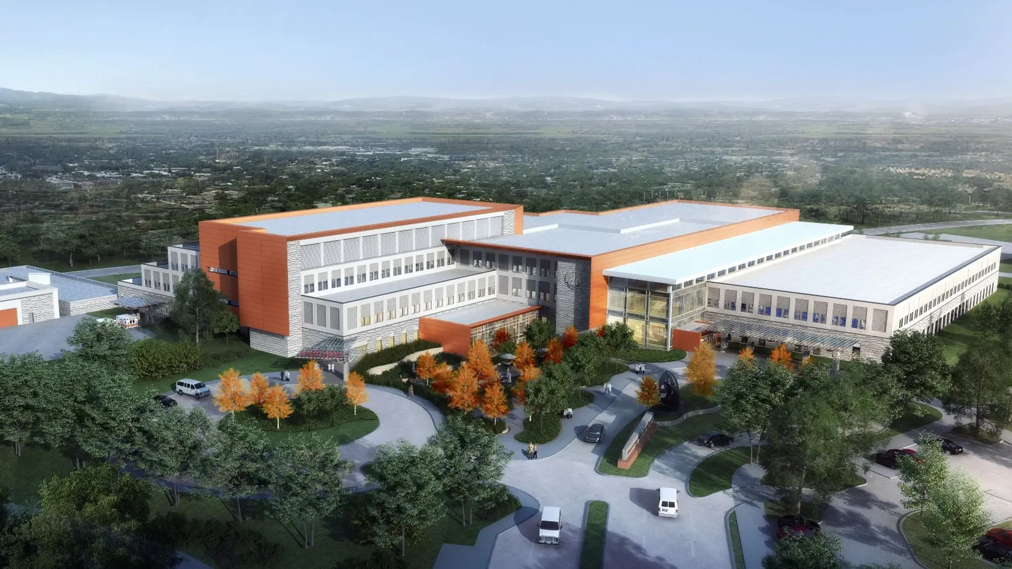 A modern multi-story building with orange accents, large windows, surrounded by trees with autumn foliage, and a parking area with several cars in front.