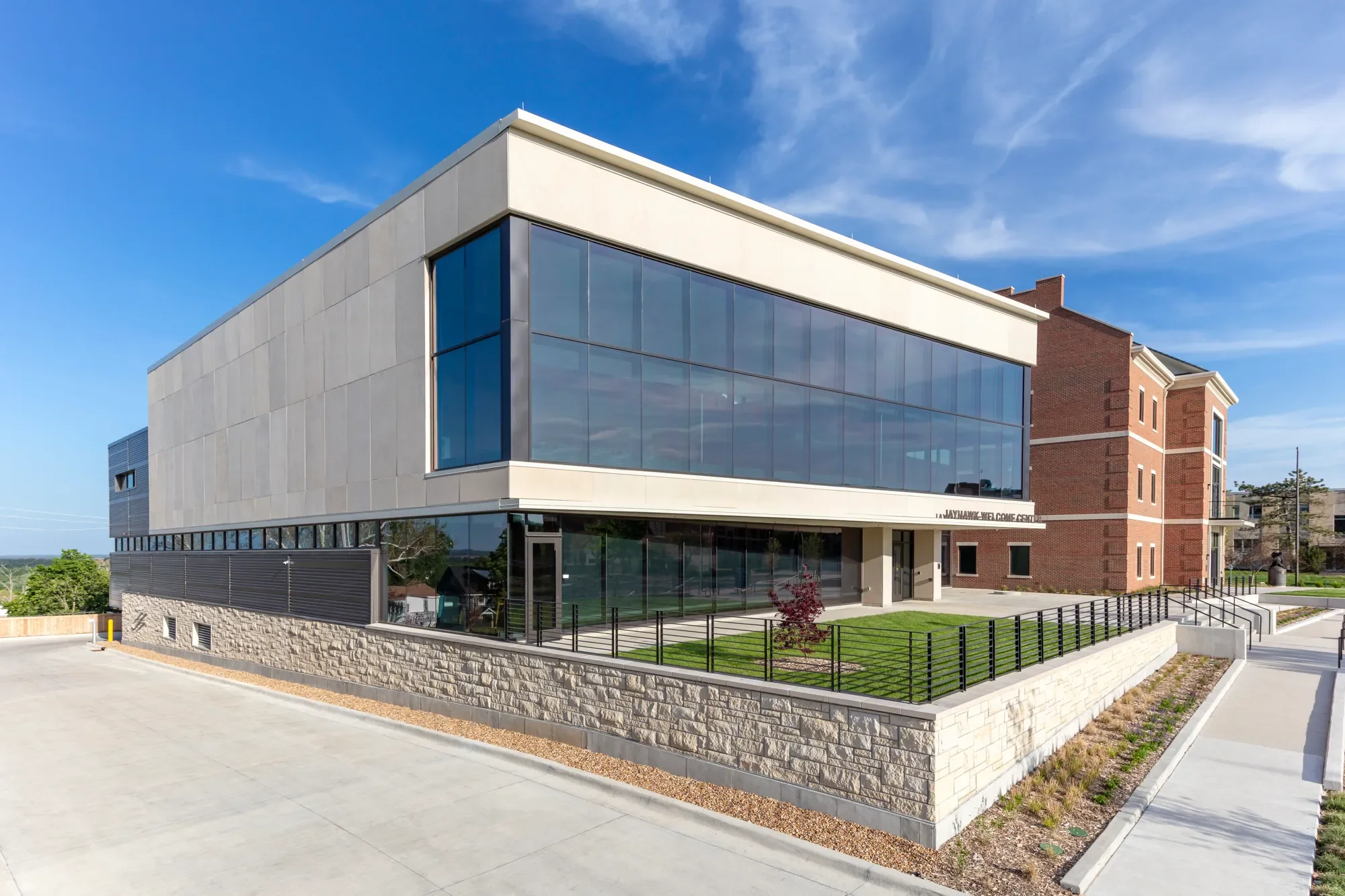 Modern building with large glass windows, gray and brick exterior, and a landscaped area with small trees, surrounded by a paved parking lot and sidewalk, under a blue sky with some clouds.