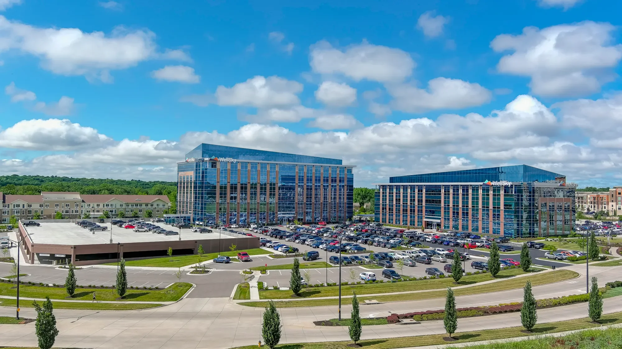 Two modern glass office buildings with the WellSky logo, surrounded by a parking lot filled with cars and a landscaped area with trees. Residential buildings are visible in the background under a partly cloudy sky.