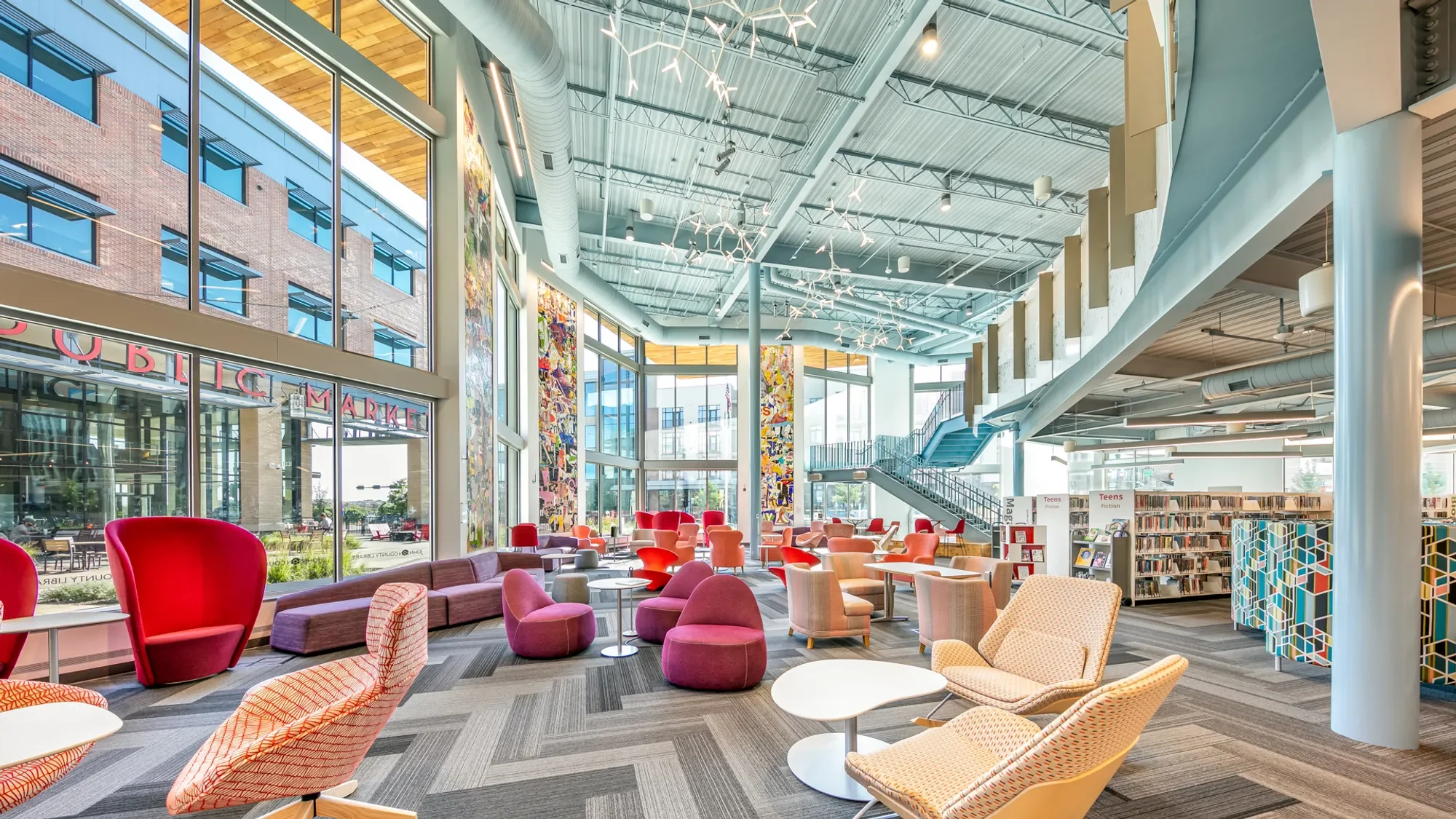 A spacious, modern indoor library with large floor-to-ceiling windows, colorful chairs, and abstract artwork on the walls. There's a staircase leading to an upper level, and the ceiling has contemporary light fixtures resembling branches.