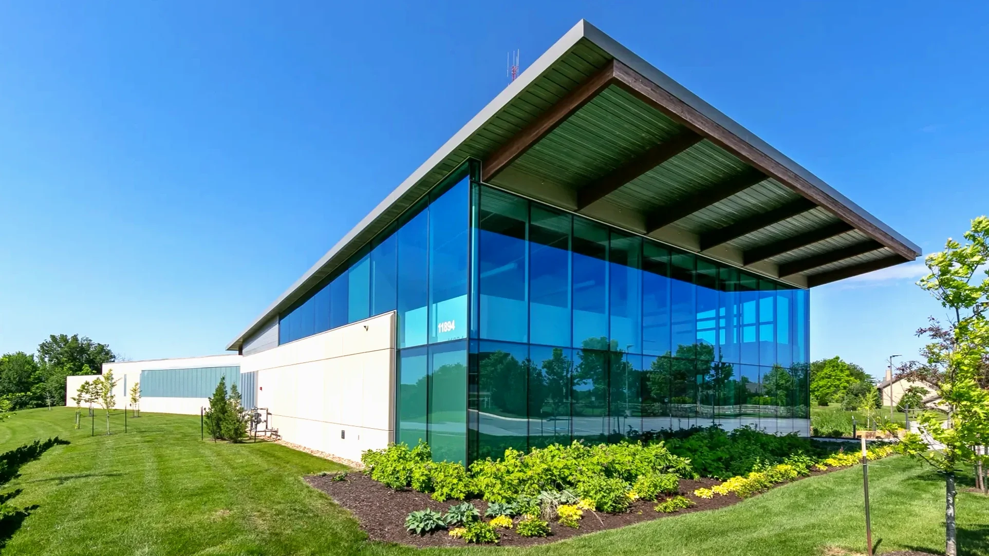 Modern commercial building with large glass windows and a flat roof, surrounded by green grass and small trees, under a clear blue sky.