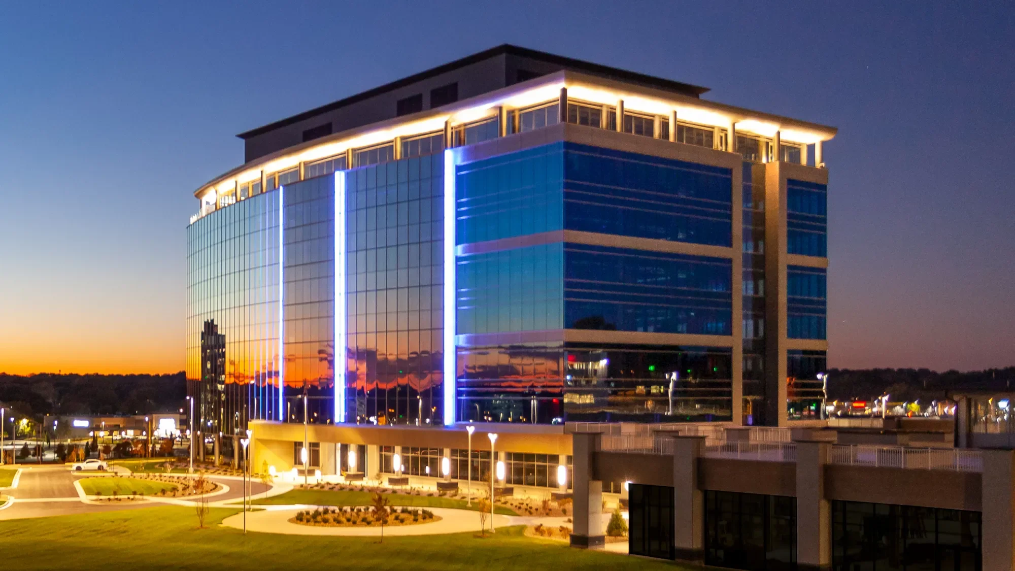 Modern glass office building illuminated with blue and white lights during sunset, with trees, grass, parking lot, and a clear sky in the background.