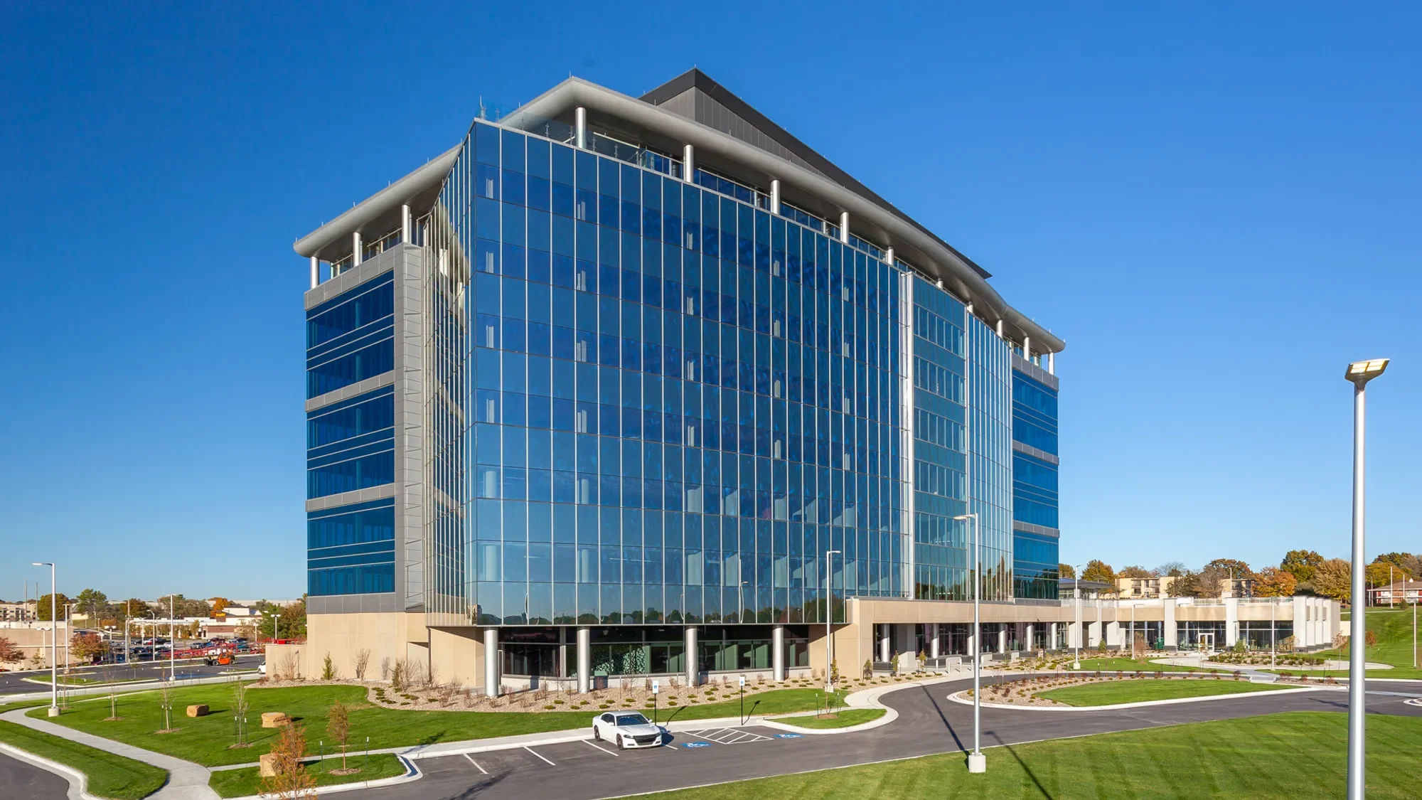 A modern office building with a glass facade reflecting the blue sky, surrounded by a parking lot, street lamps, and landscaped green areas with trees.