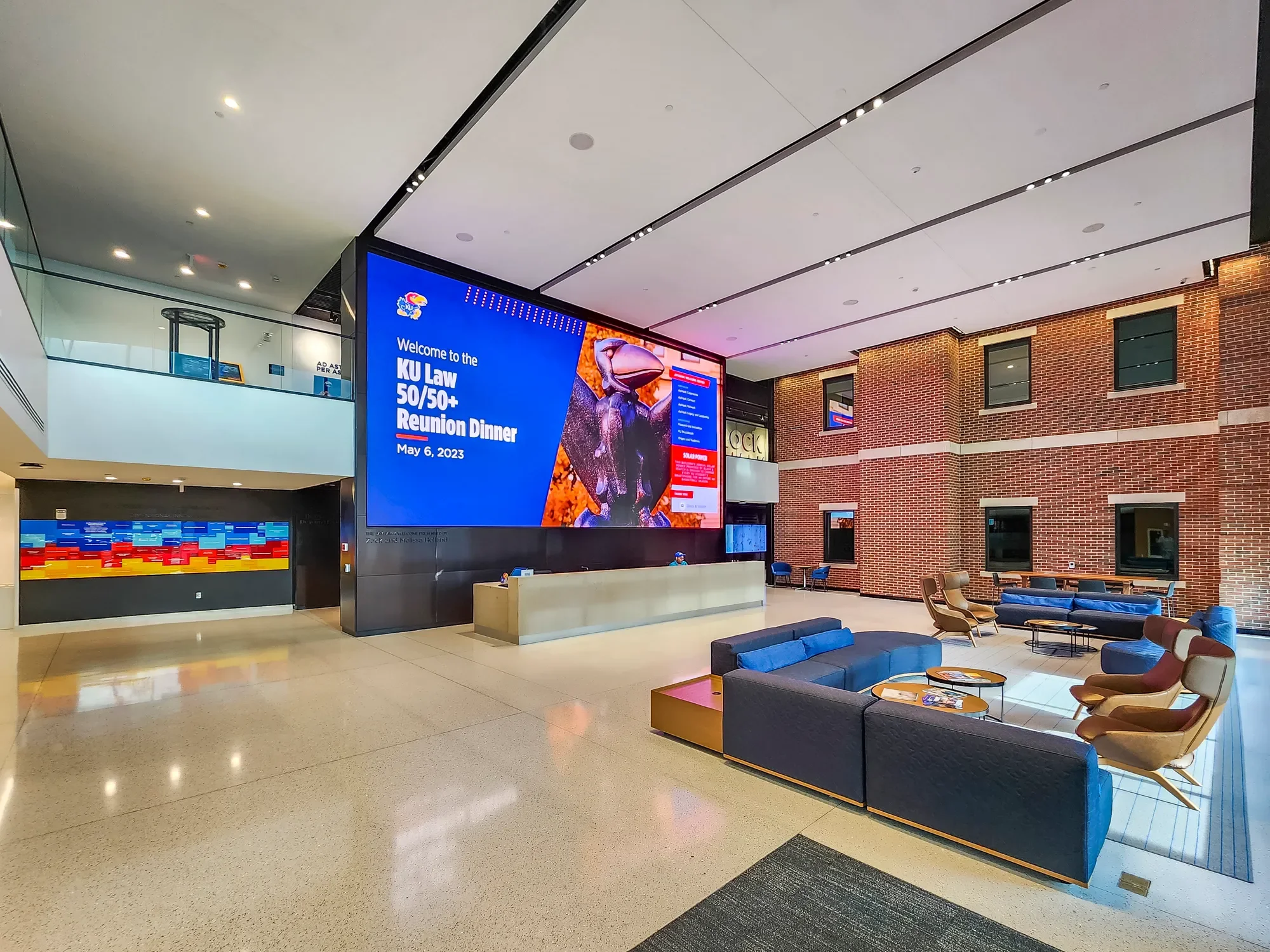 Inside of a modern airport terminal with seating areas, a large digital screen displaying an announcement for a KU Law reunion dinner on May 6, 2023, brick walls, and a high ceiling with lighting.