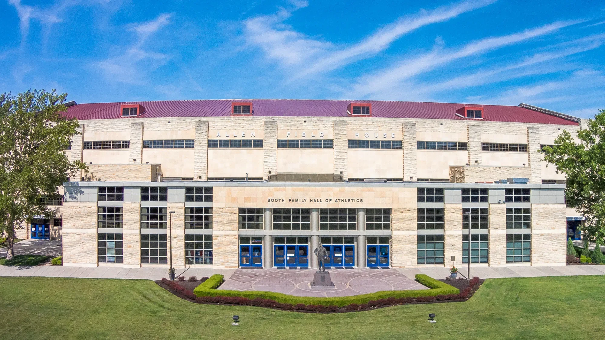 Front view of Allen Field House, a large sports arena with stone exterior, blue doors, windows, a small statue in front, and trees on either side under a partly cloudy sky.