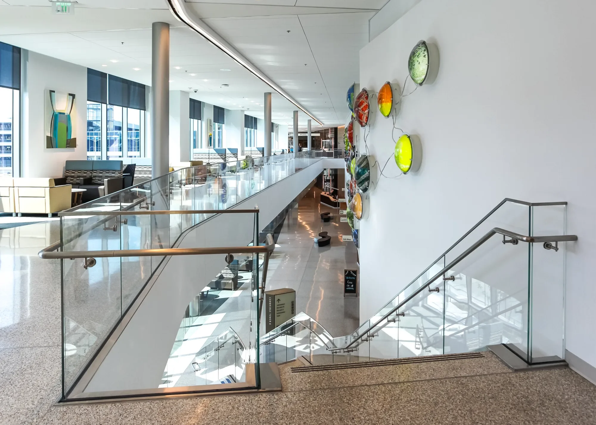 Interior of a modern building with large windows, seating area, colorful decorative glass art on the wall, and a staircase with glass railings leading down.