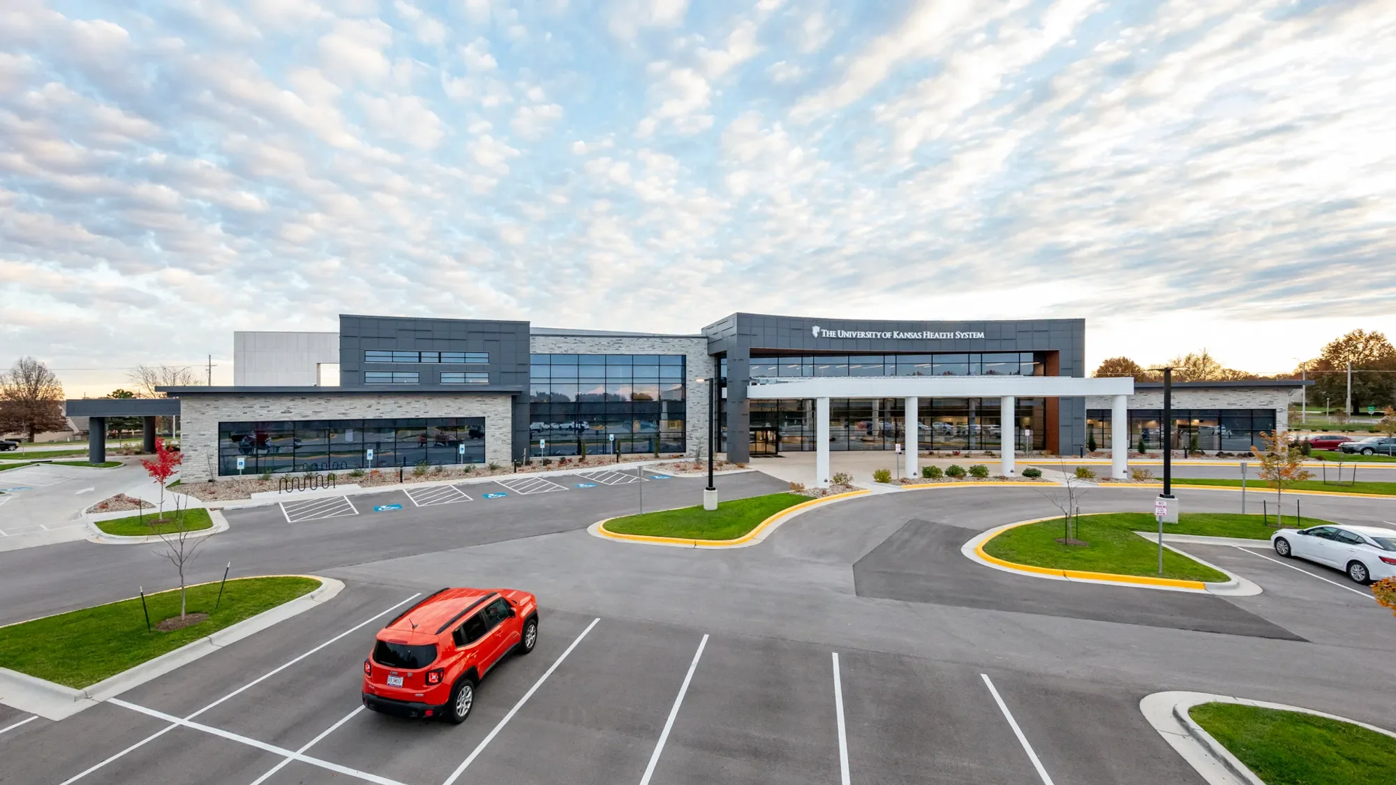 Modern building with a sign reading 'The University of Kansas Health System.' In the foreground, there is a parking lot with several cars, including a red SUV, and neatly landscaped areas with small trees and grass.