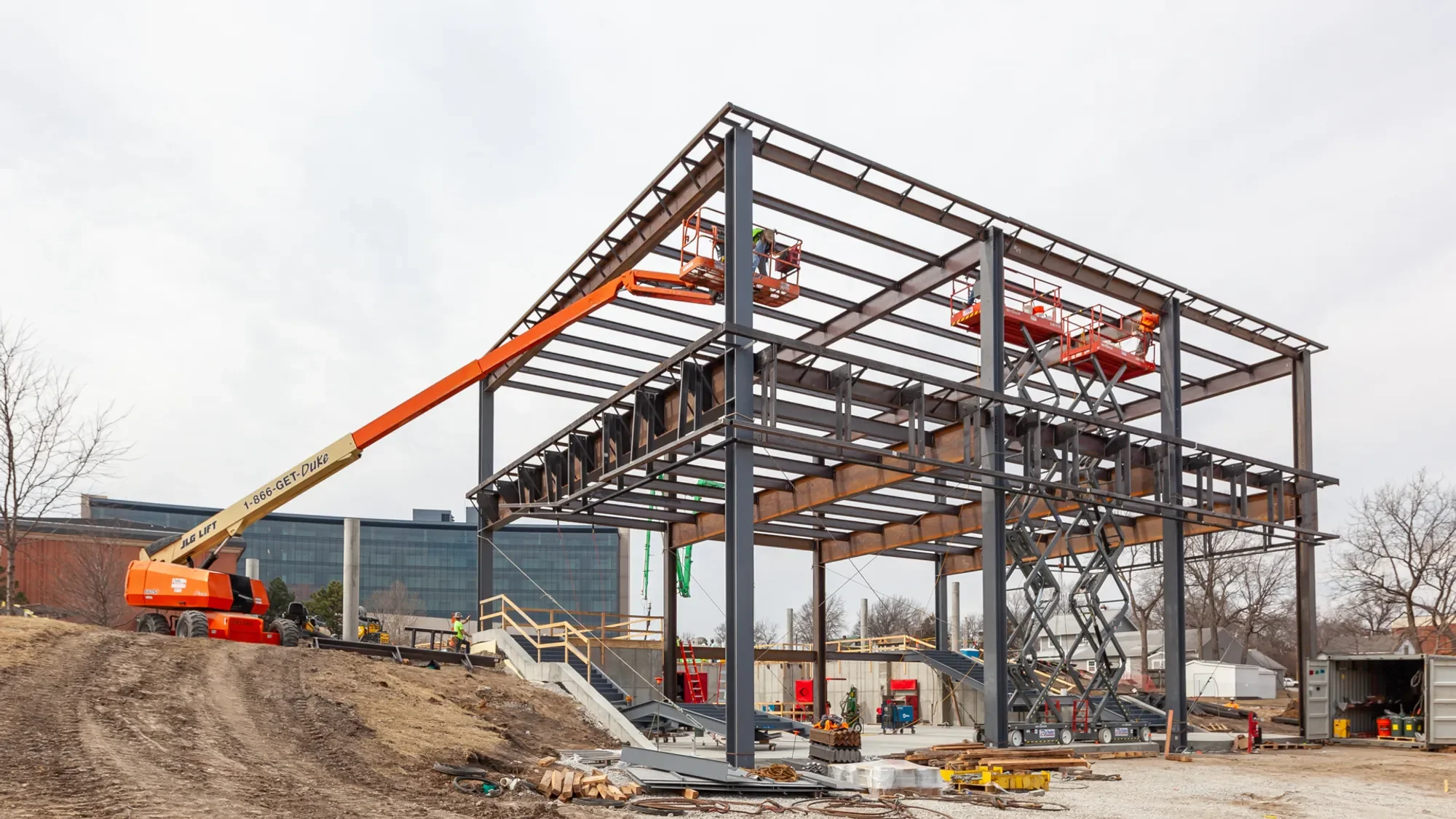 Construction site with a steel building framework in progress, workers, and an orange crane lift.