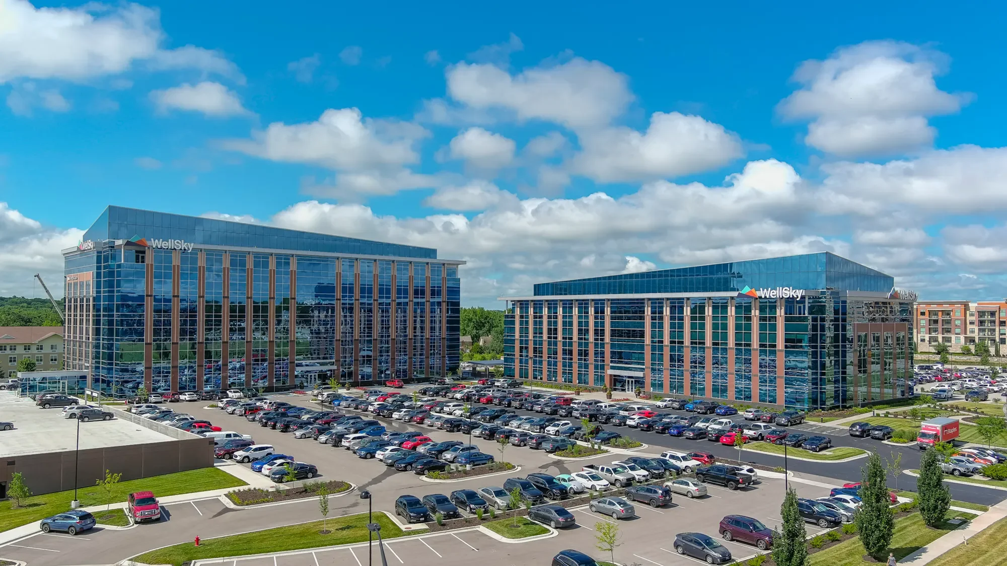 Two modern office buildings with glass facades, labeled WellSky, surrounded by a parking lot filled with cars and additional buildings in the background under a partly cloudy blue sky.