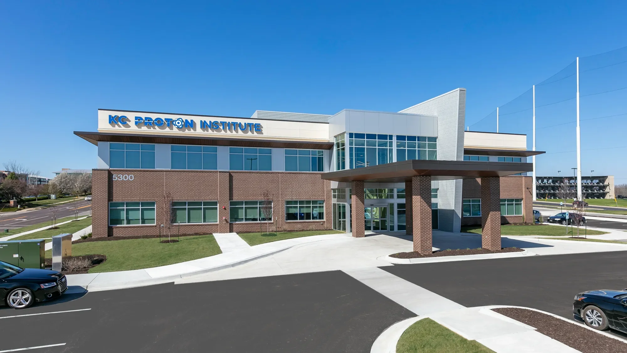 Modern educational building with the sign 'KC Proton Institute' on the front, surrounded by parking lot, grassy areas, and clear sky.
